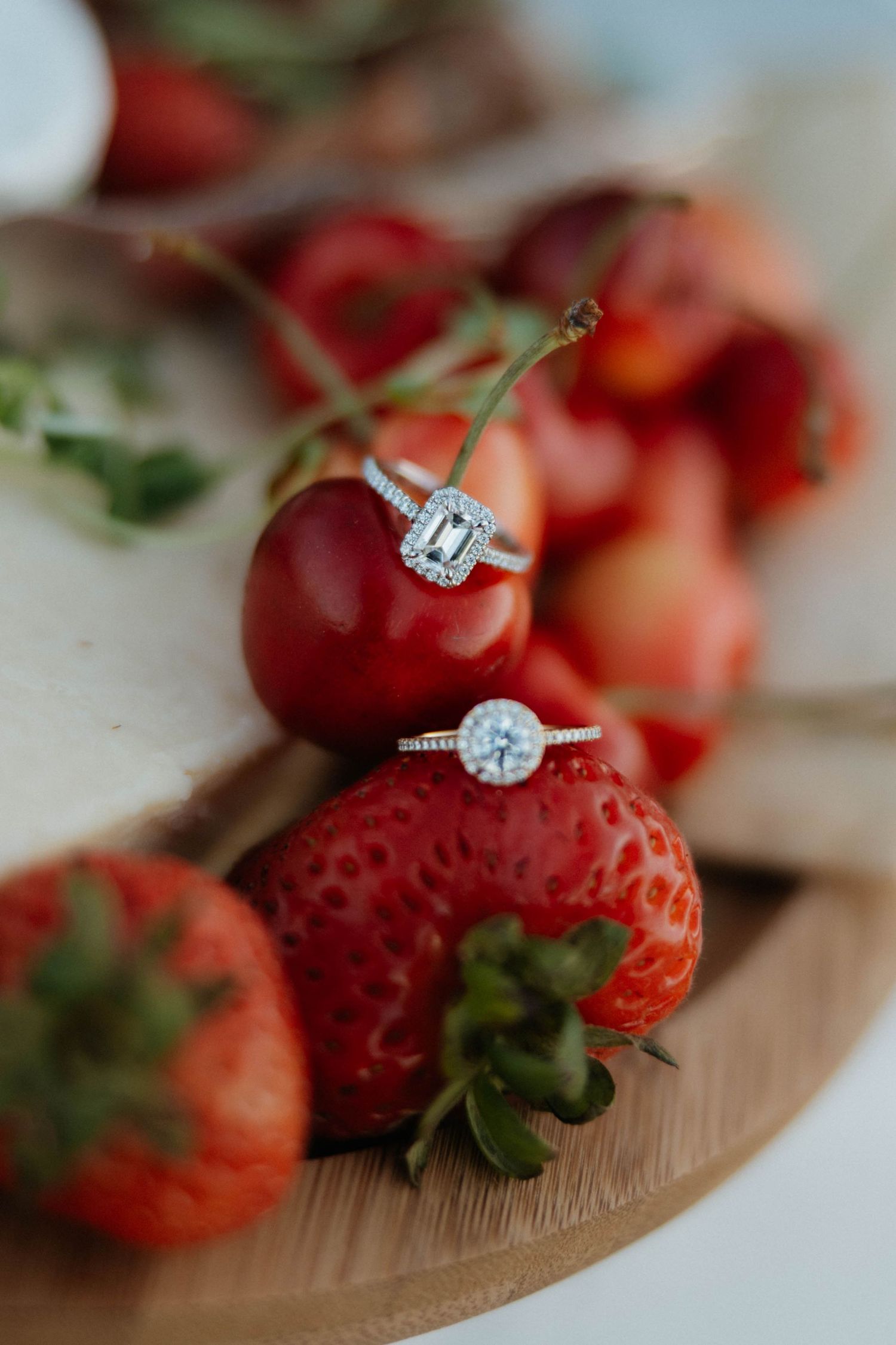 Two diamond halo engagement rings resting on fresh strawberries and cherries on a wooden board.