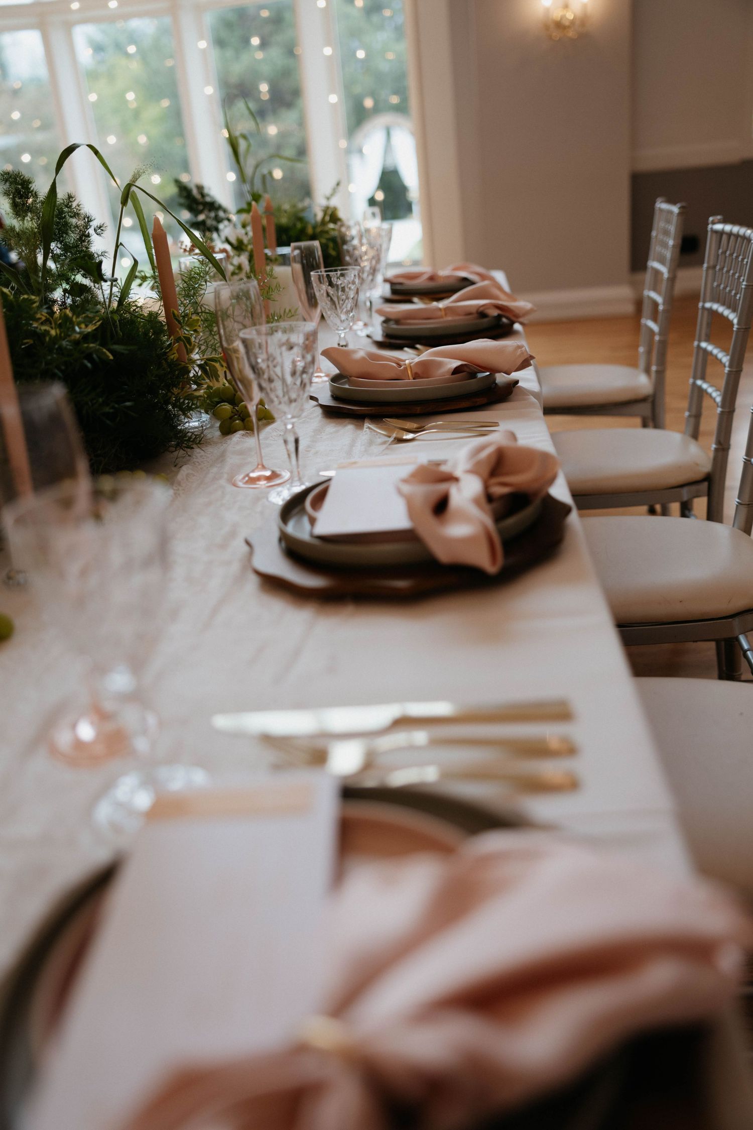 Elegant wedding reception table with blush pink napkins, gold cutlery, dark plates, and a lush greenery centerpiece runner.