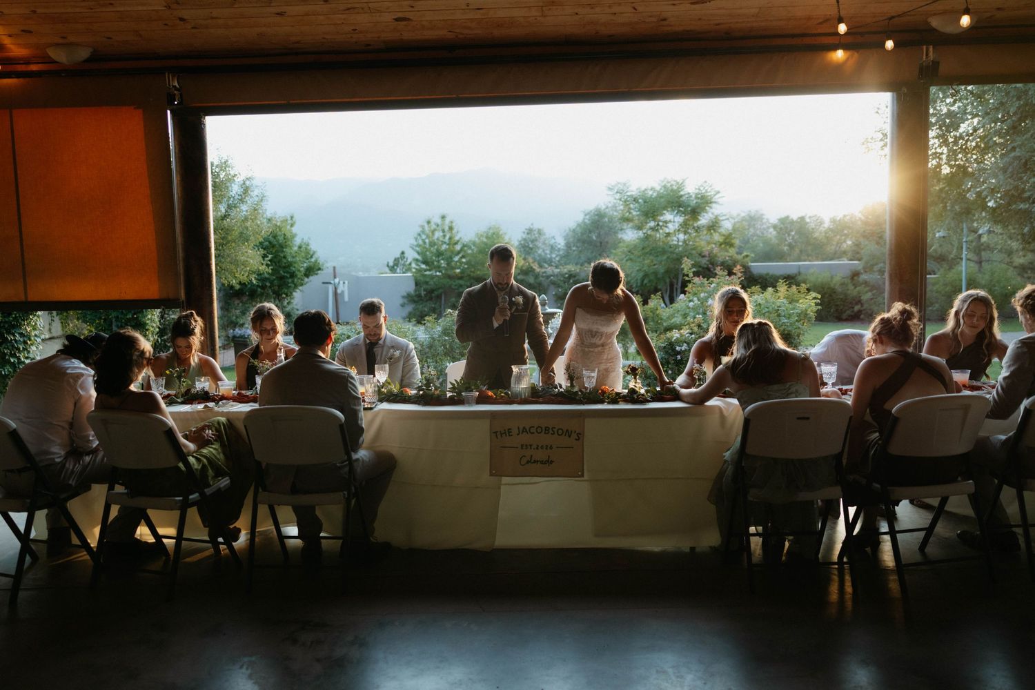 Wedding reception dinner with guests seated at long tables, backlit by golden sunset mountain views through open venue walls.