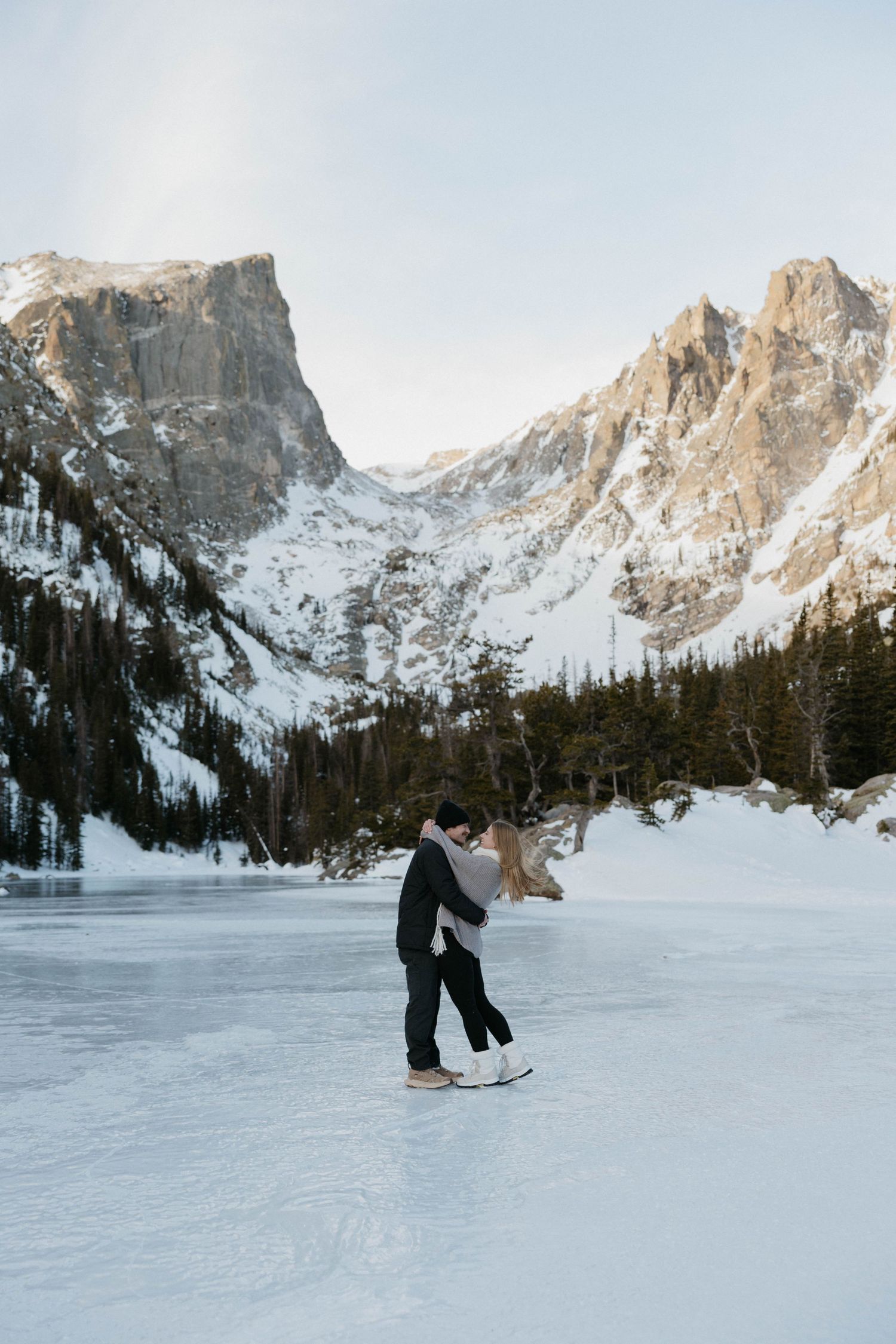 Couple ice skating on a frozen mountain lake surrounded by snow-capped peaks and pine trees in winter.