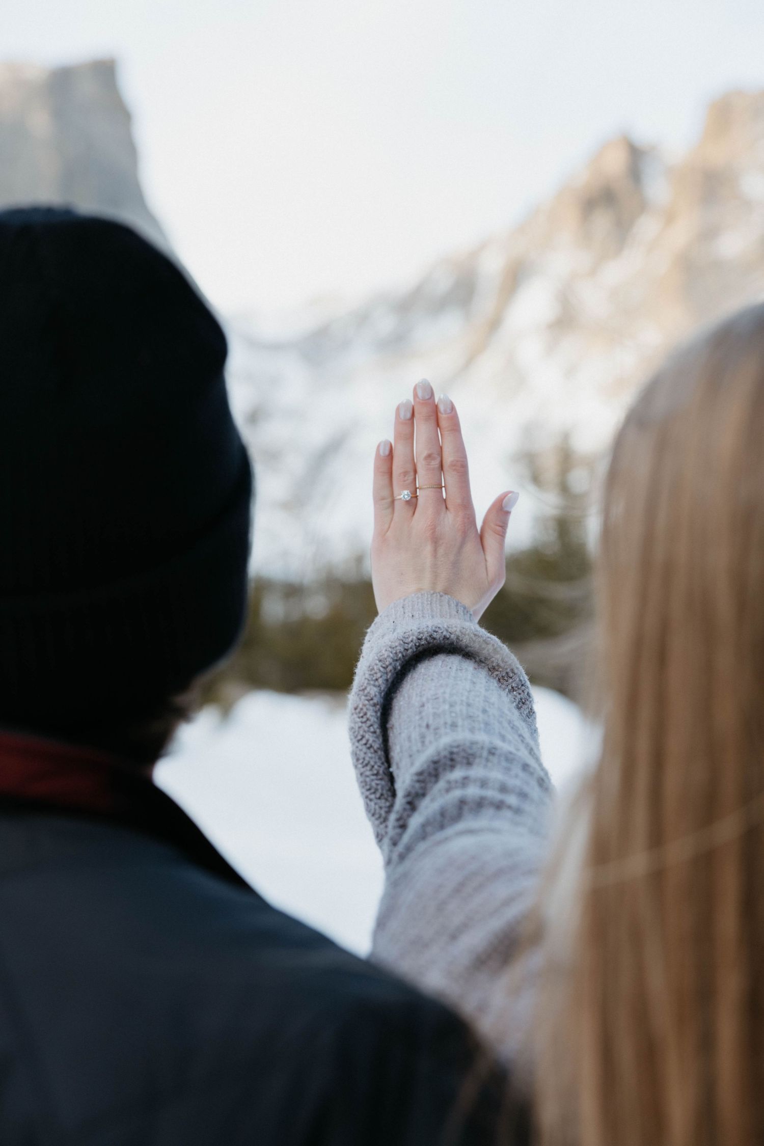 Couple in winter mountains, woman showing engagement ring with snowy peaks in background.
