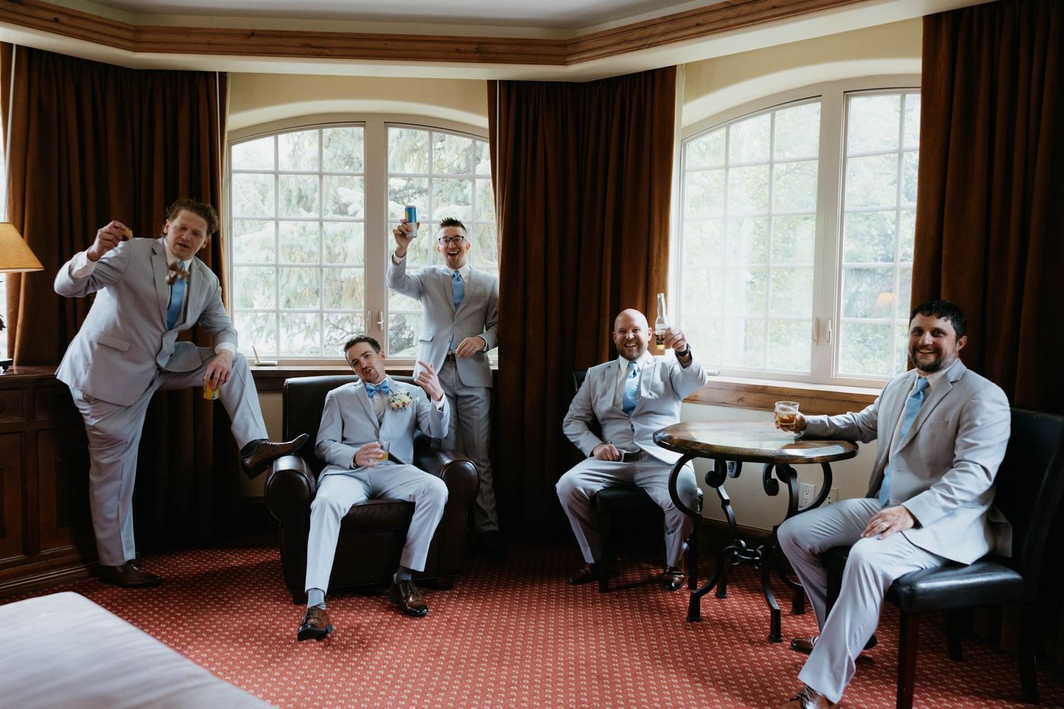 Groomsmen in light gray suits celebrate together in an elegant hotel room, raising glasses in a joyful pre-wedding toast.