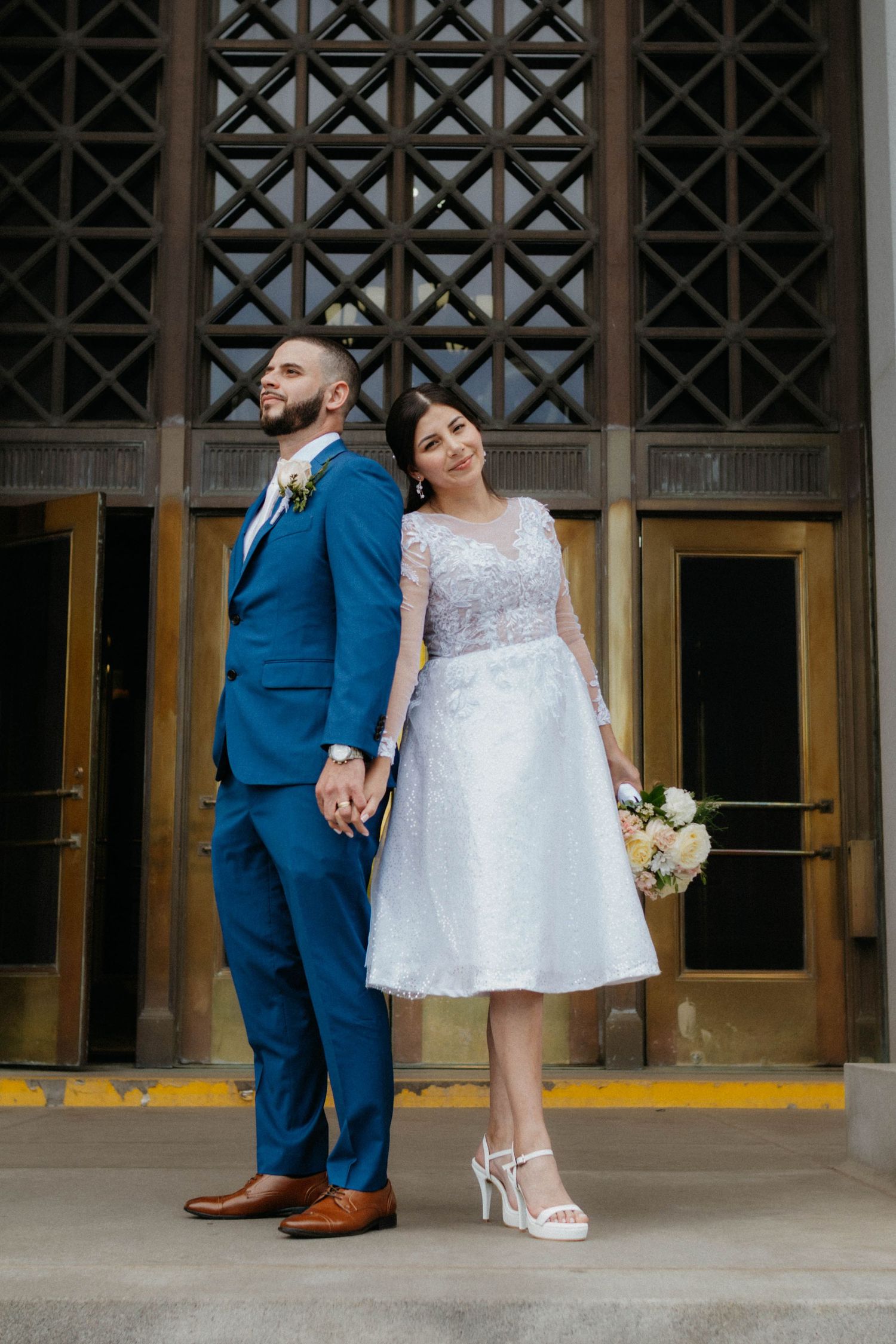 Bride in short white lace dress and groom in blue suit hold hands posing in front of ornate building doors.