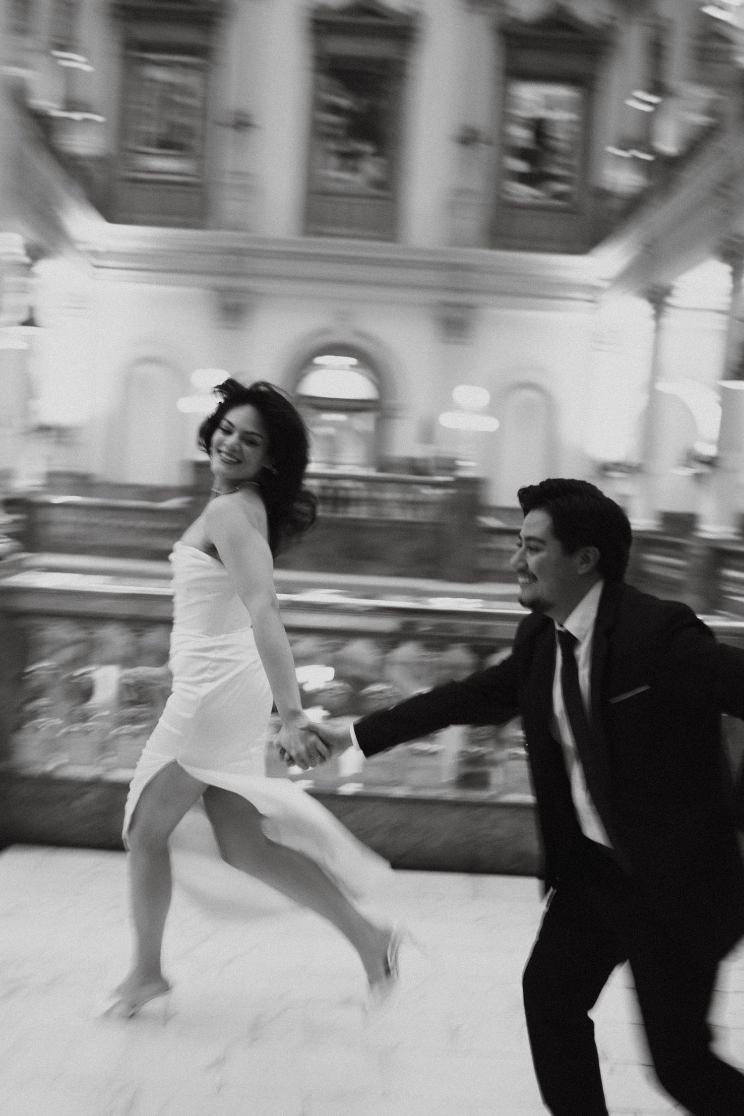 Black and white photo of a couple in formal attire joyfully running hand in hand inside an elegant venue.