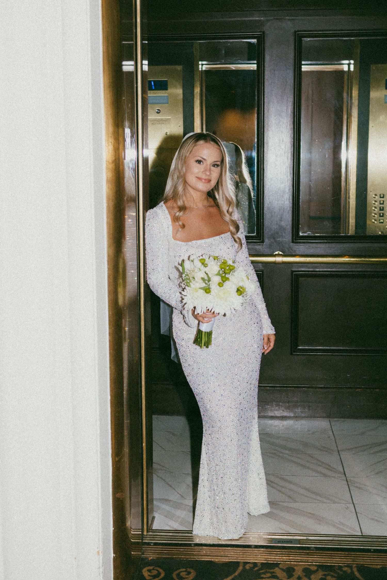 A bride in a white lace wedding dress holds a bouquet while posing in an elegant indoor setting.