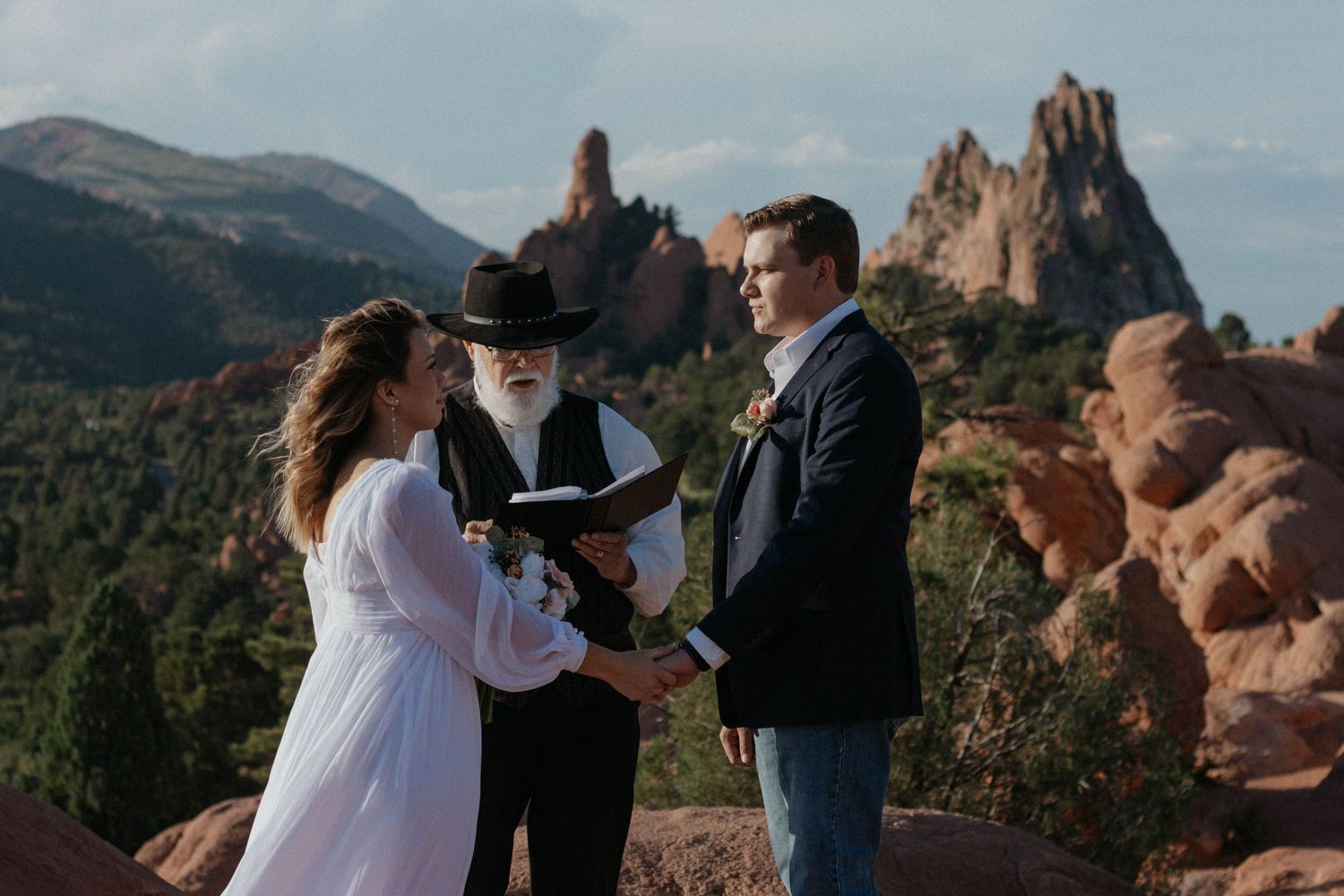 Couple exchanges vows at Garden of the Gods, Colorado, officiated by a bearded man amid red rock formations.