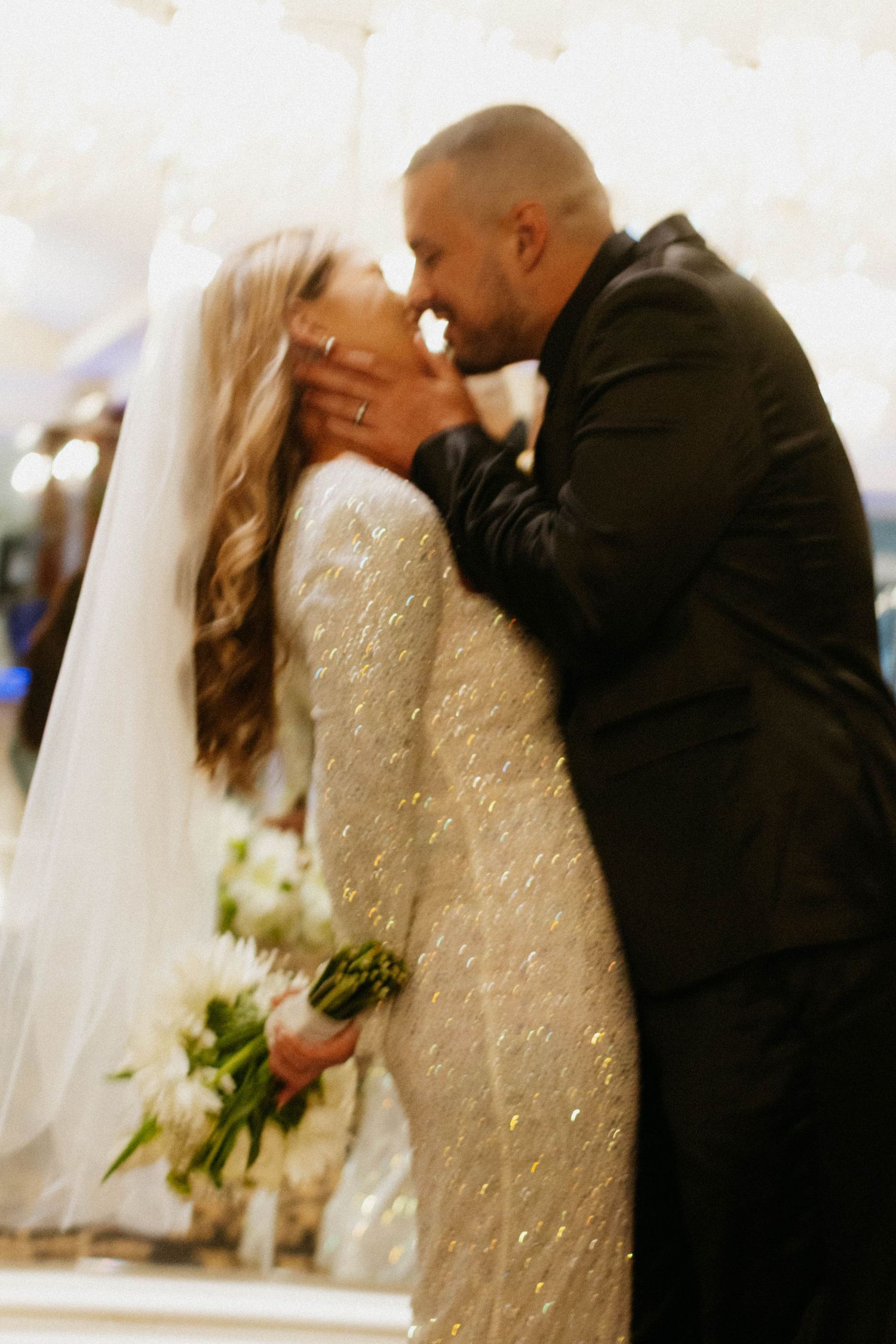A bride in a sparkly wedding gown dips back for a romantic kiss with her groom during their wedding celebration.