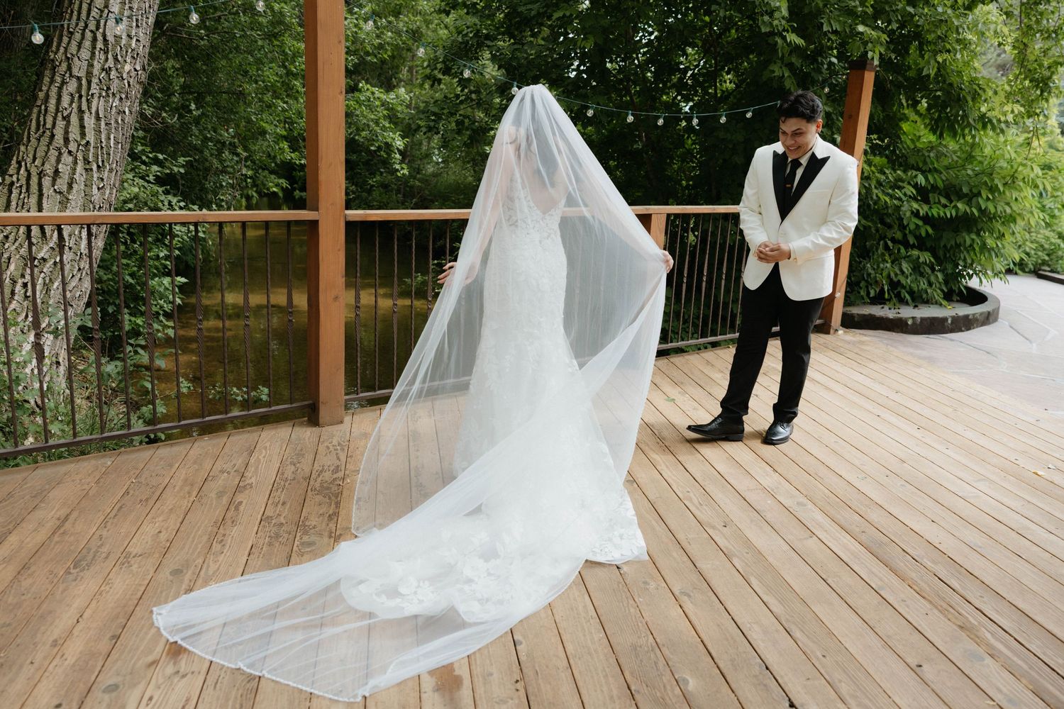 Bride in elegant gown with long veil stands with groom on wooden deck surrounded by lush greenery.