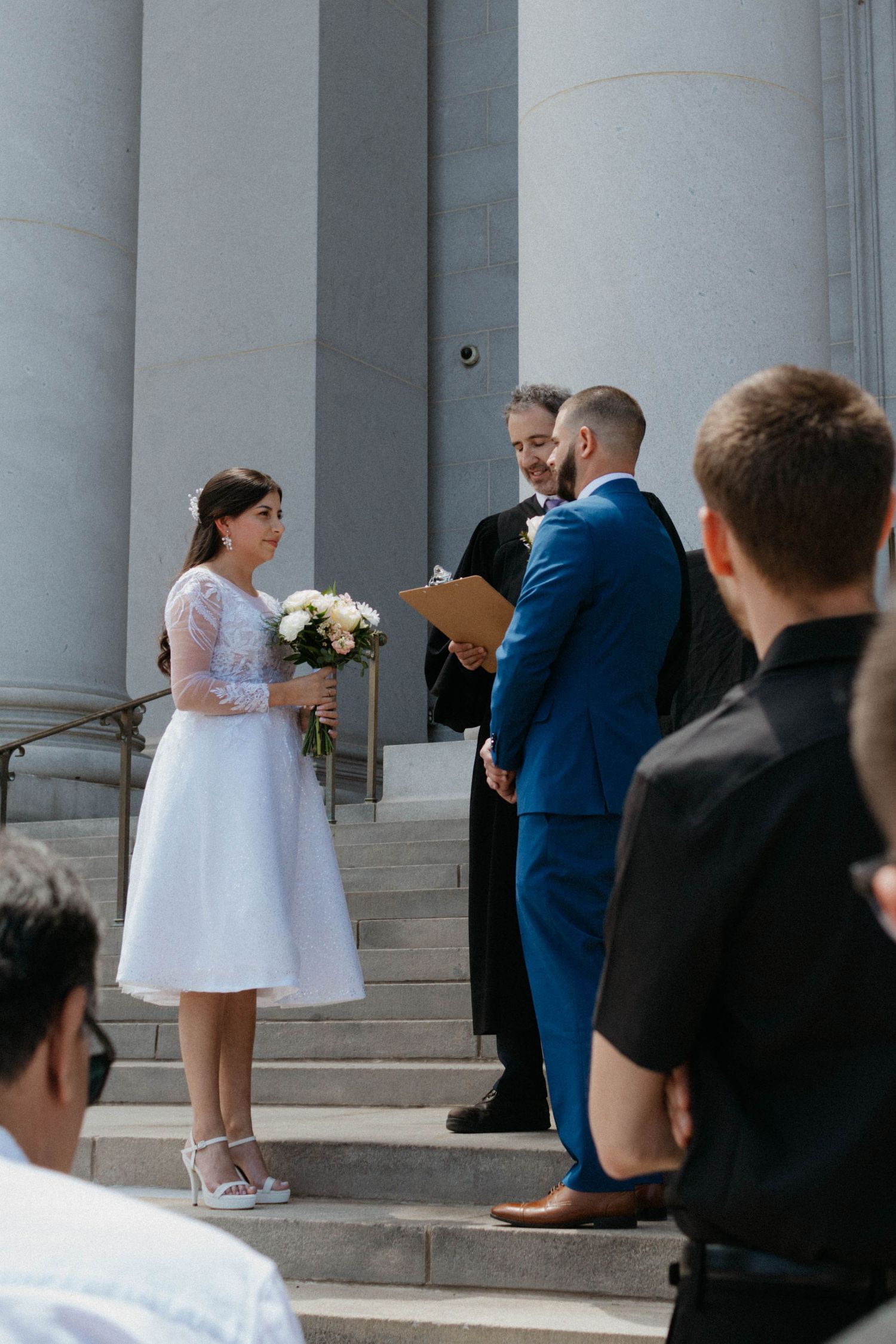 Bride and groom exchanging vows on stone steps with columns, officiant holding clipboard, guests watching outdoor ceremony.