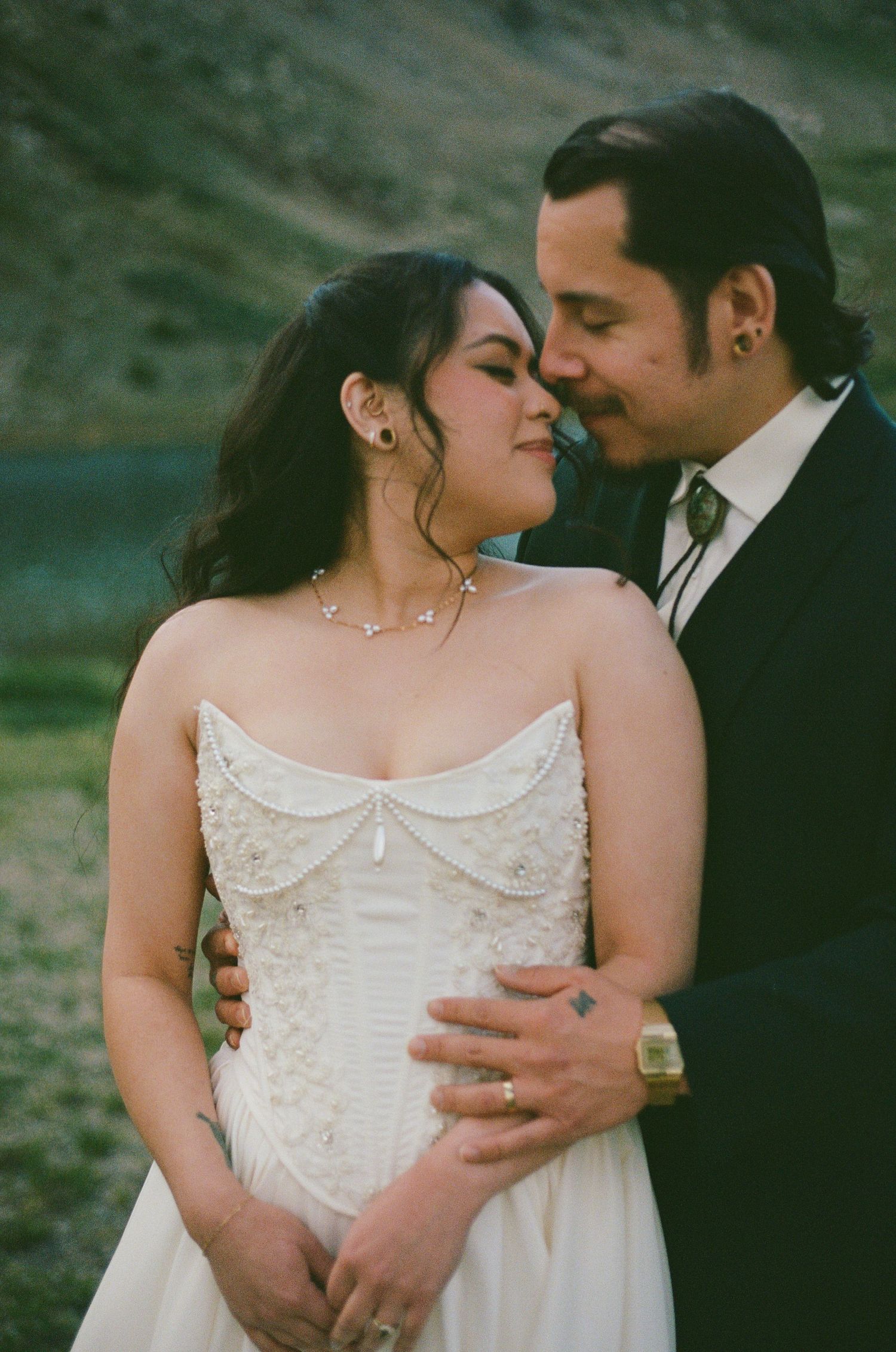 A bride in a strapless embroidered gown and groom in a dark suit share a tender moment outdoors near a mountain lake.