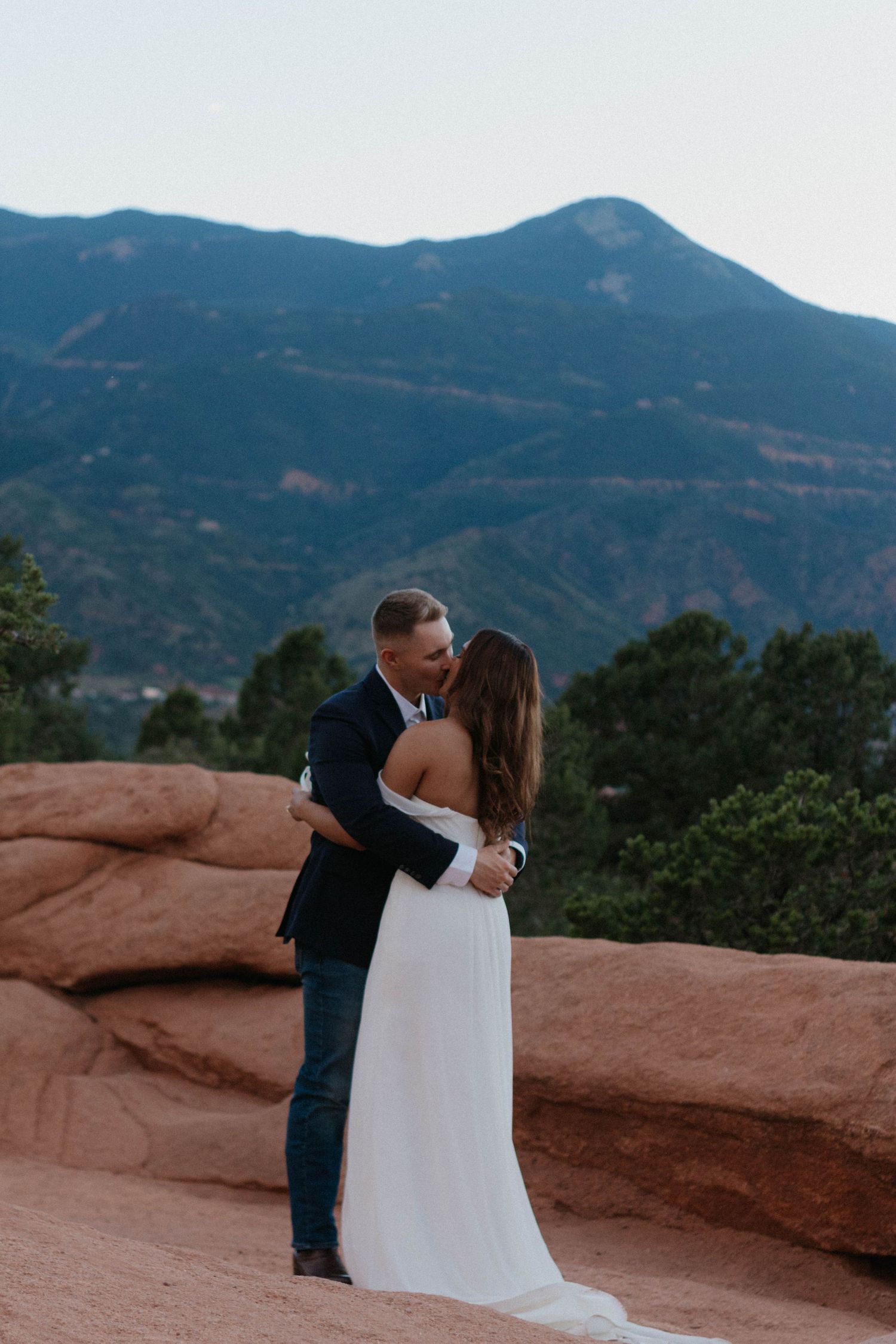 Couple sharing a kiss in wedding attire at Garden of the Gods with red rocks and mountain backdrop.