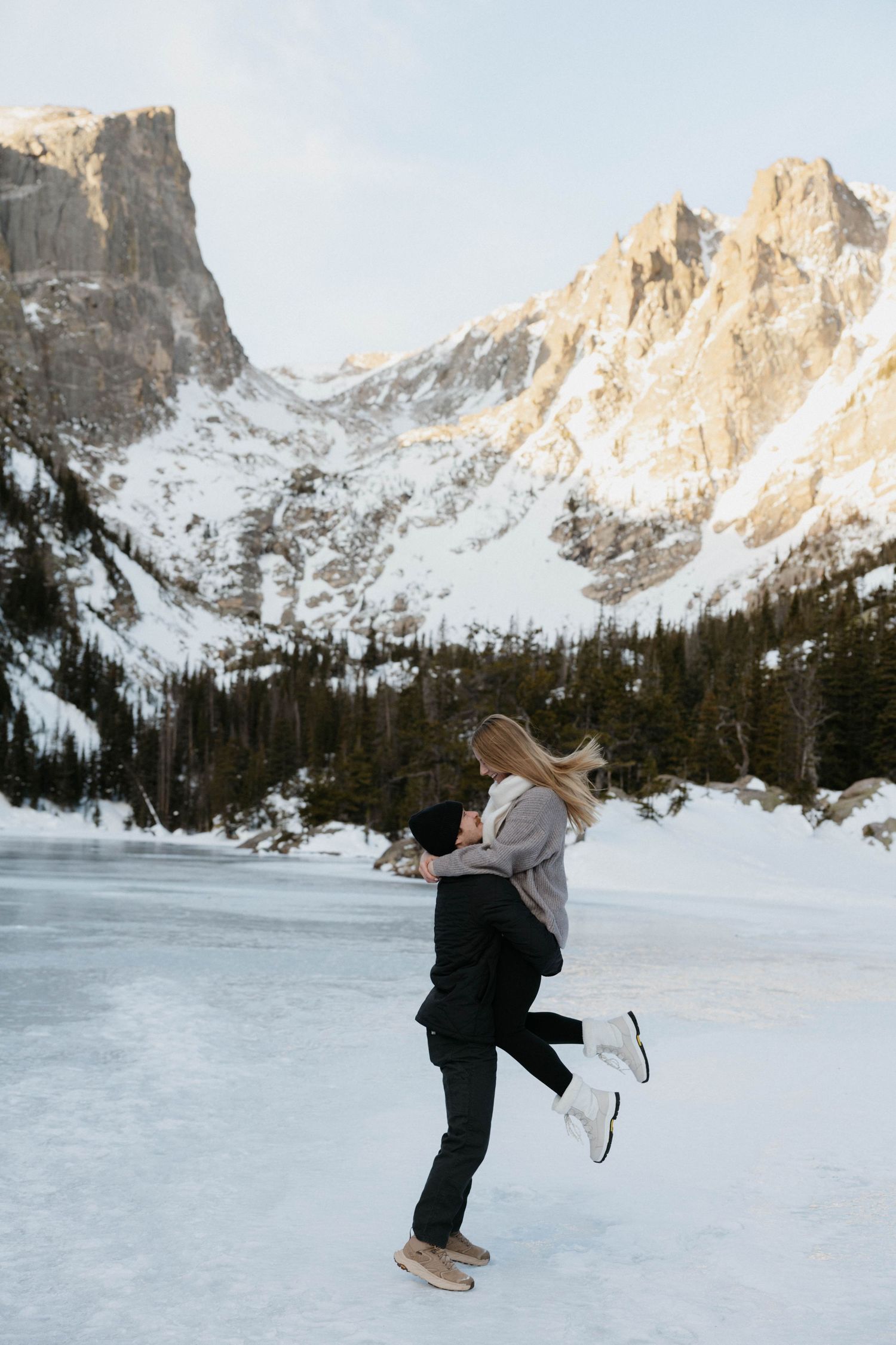 Couple embracing on a frozen lake with snow-capped Rocky Mountain peaks and pine trees in the background.