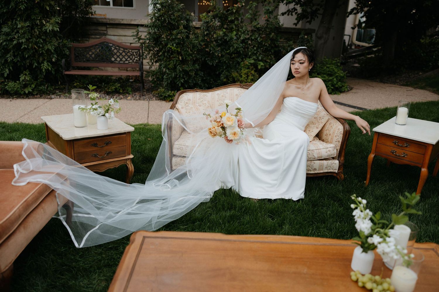 Bride in white strapless gown with flowing veil reclines on vintage sofa outdoors, holding pastel bouquet.