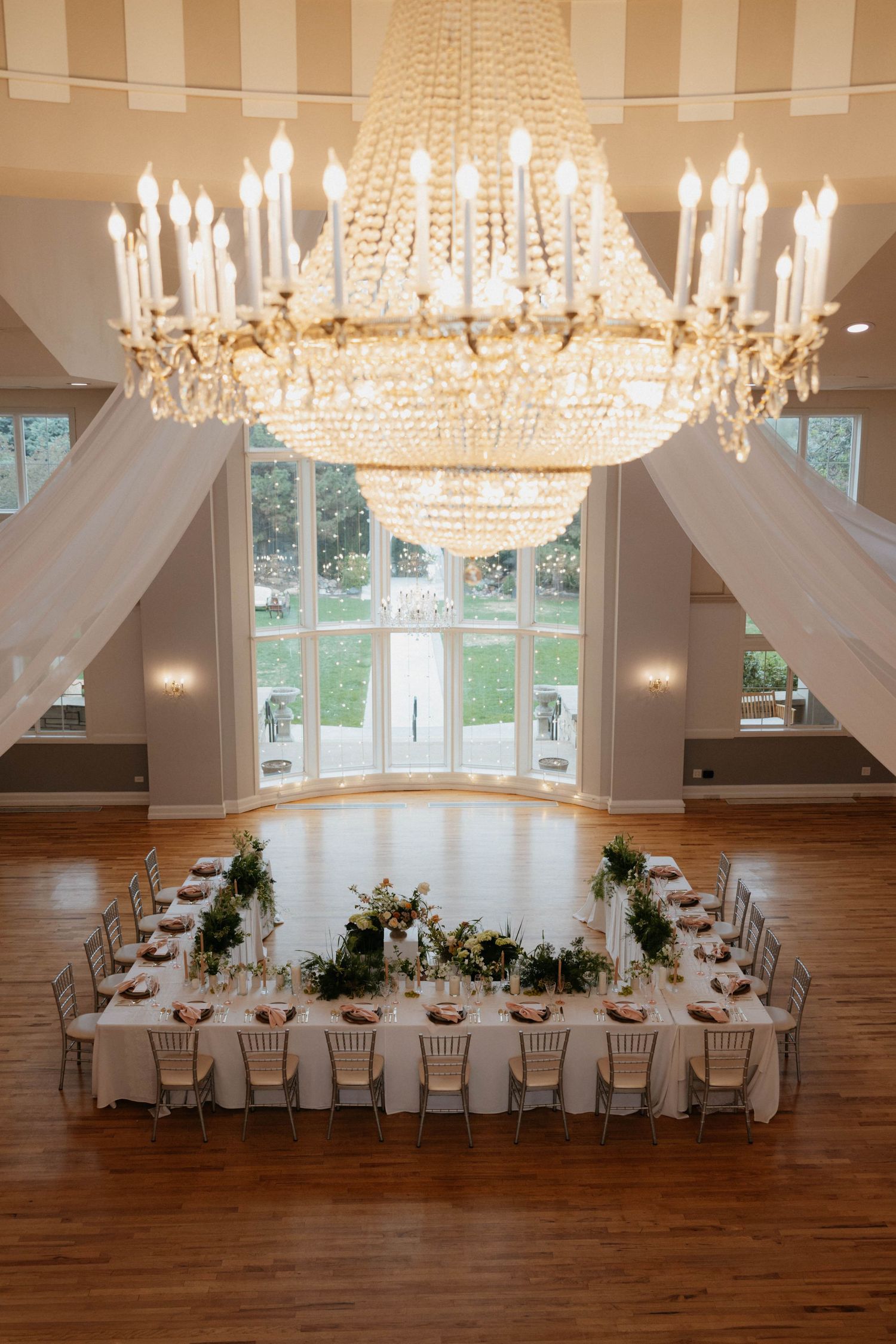 Elegant wedding reception hall with crystal chandelier, U-shaped dining table, floral centerpieces, and large windows.