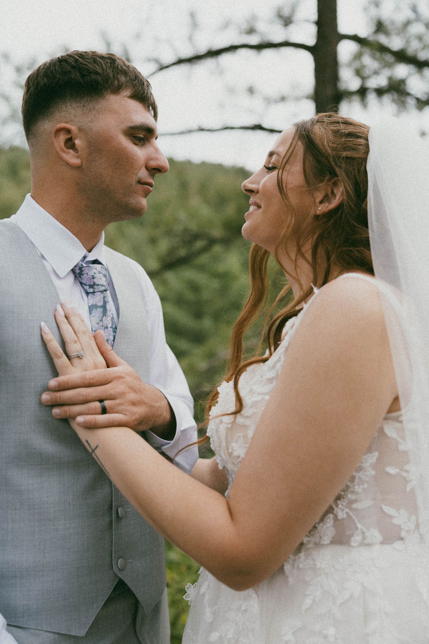 A bride and groom share a romantic moment outdoors, with the bride in lace gown and veil, groom in gray suit.