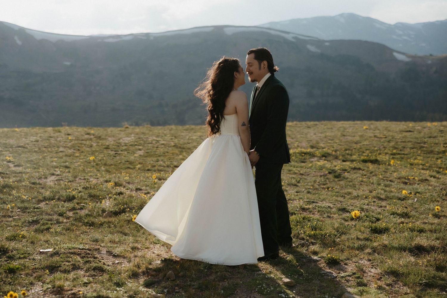 A bride and groom share a romantic moment in a scenic mountain meadow, with rolling hills and misty peaks in the background.