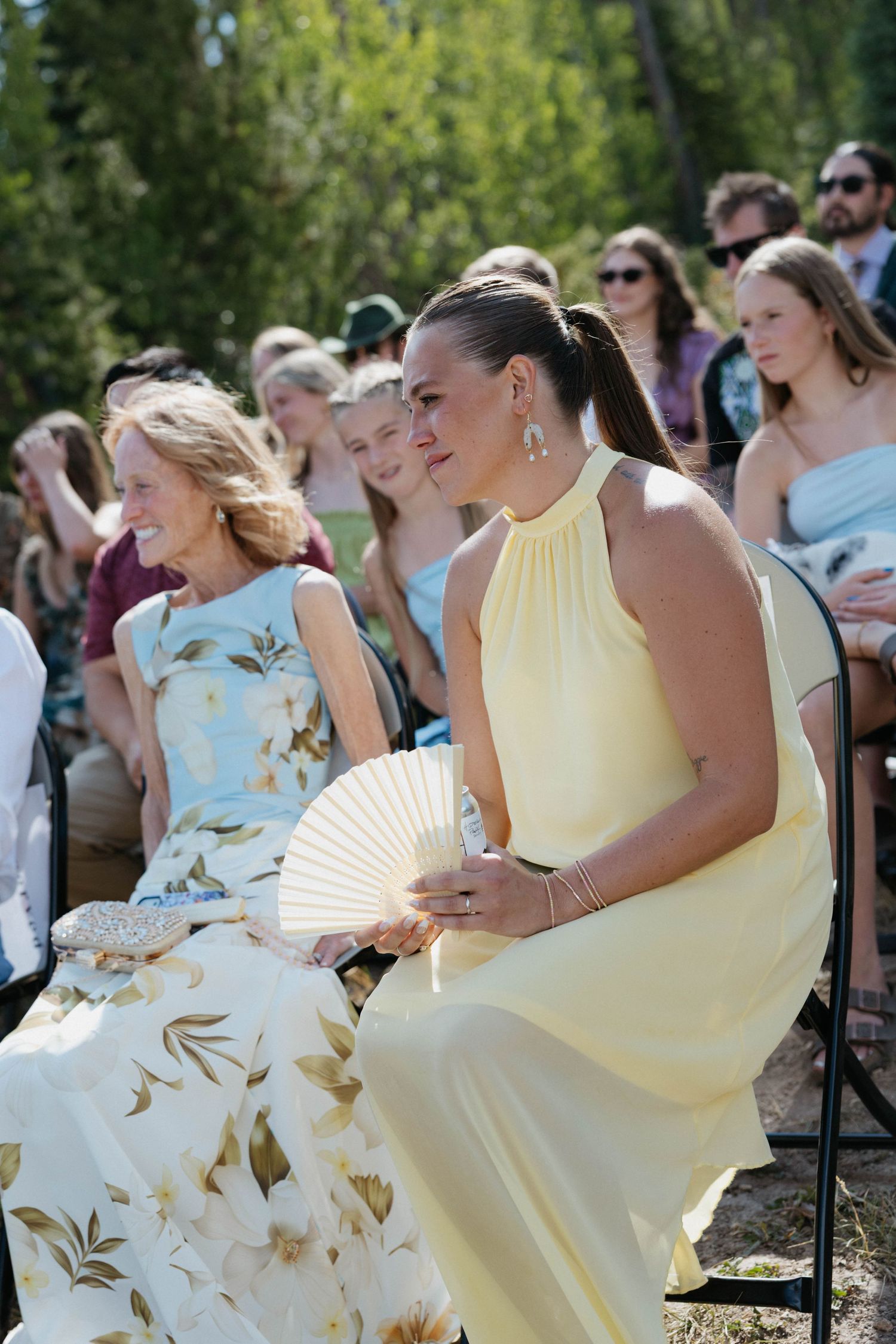 Woman in yellow dress holding fan at outdoor wedding ceremony with guests seated in background.