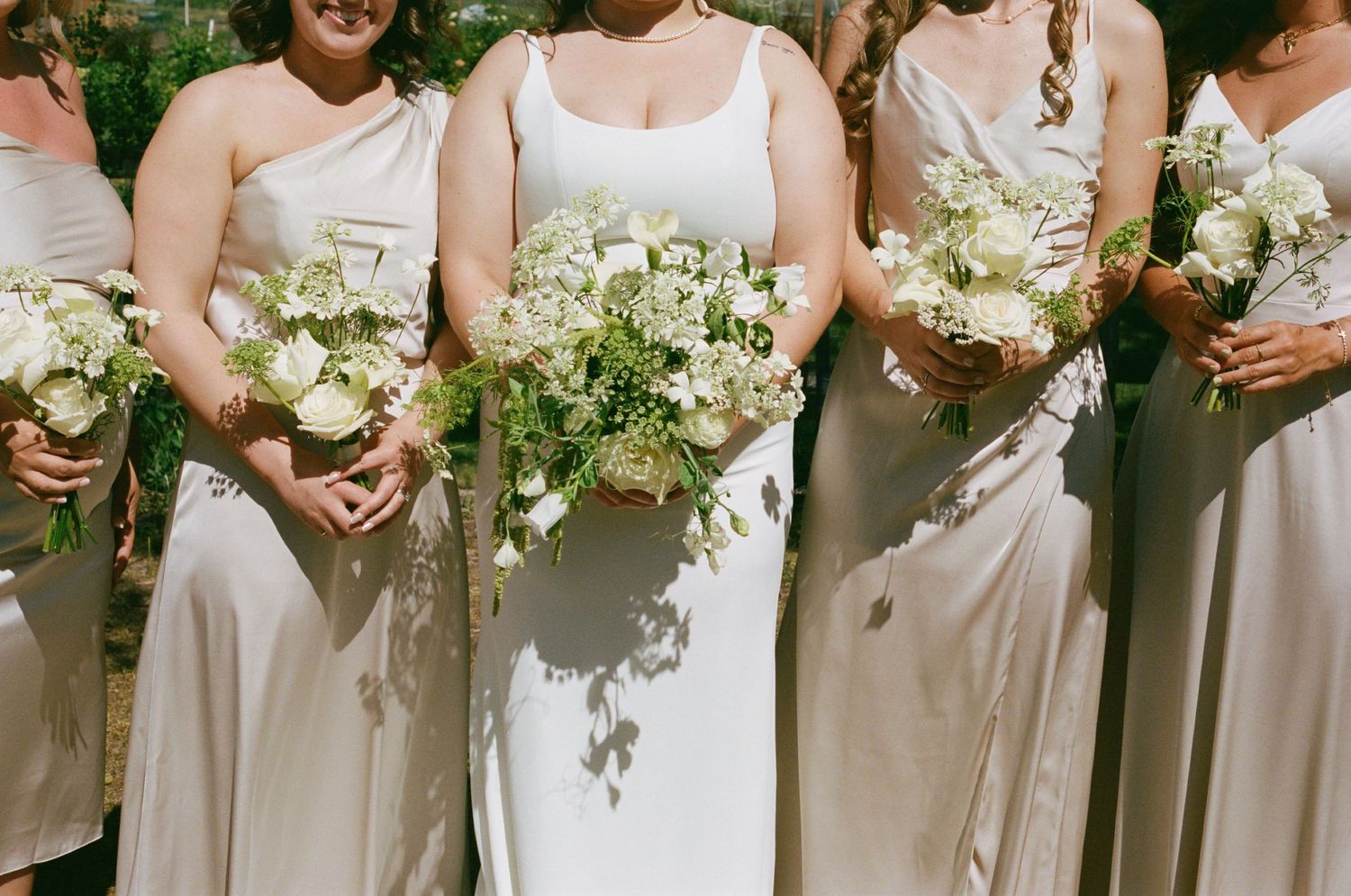 Bride and bridesmaids in ivory gowns holding lush white and green bouquets at an outdoor wedding ceremony.