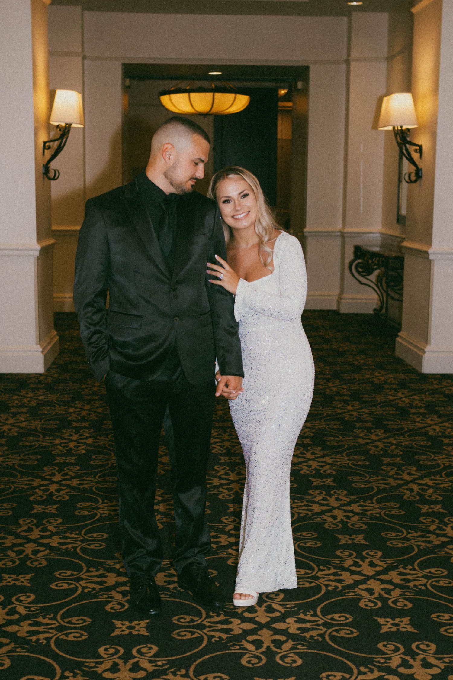 A couple poses in formal attire in an elegant hotel hallway with ornate carpet and wall sconces.