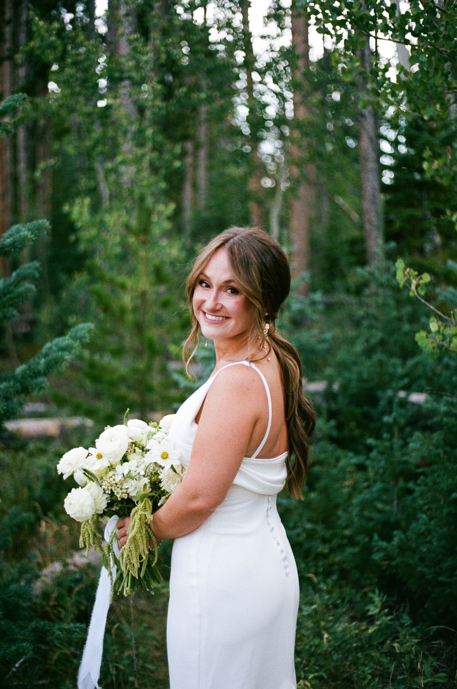 Smiling bride in white gown holding white bouquet, posing in lush green forest setting.