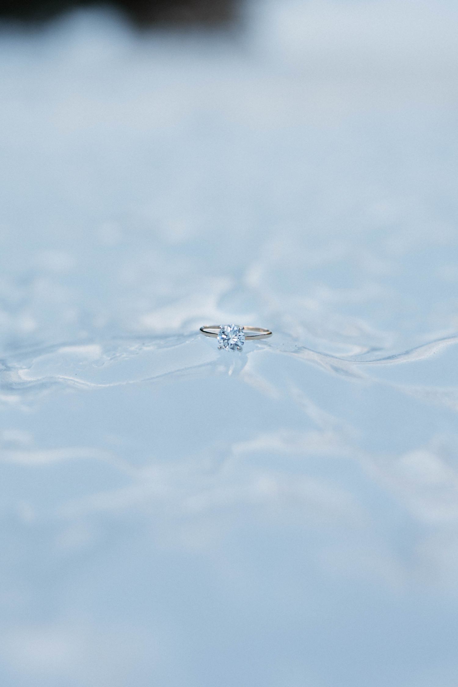 A delicate solitaire diamond engagement ring resting on a shimmering water surface with soft blue tones.