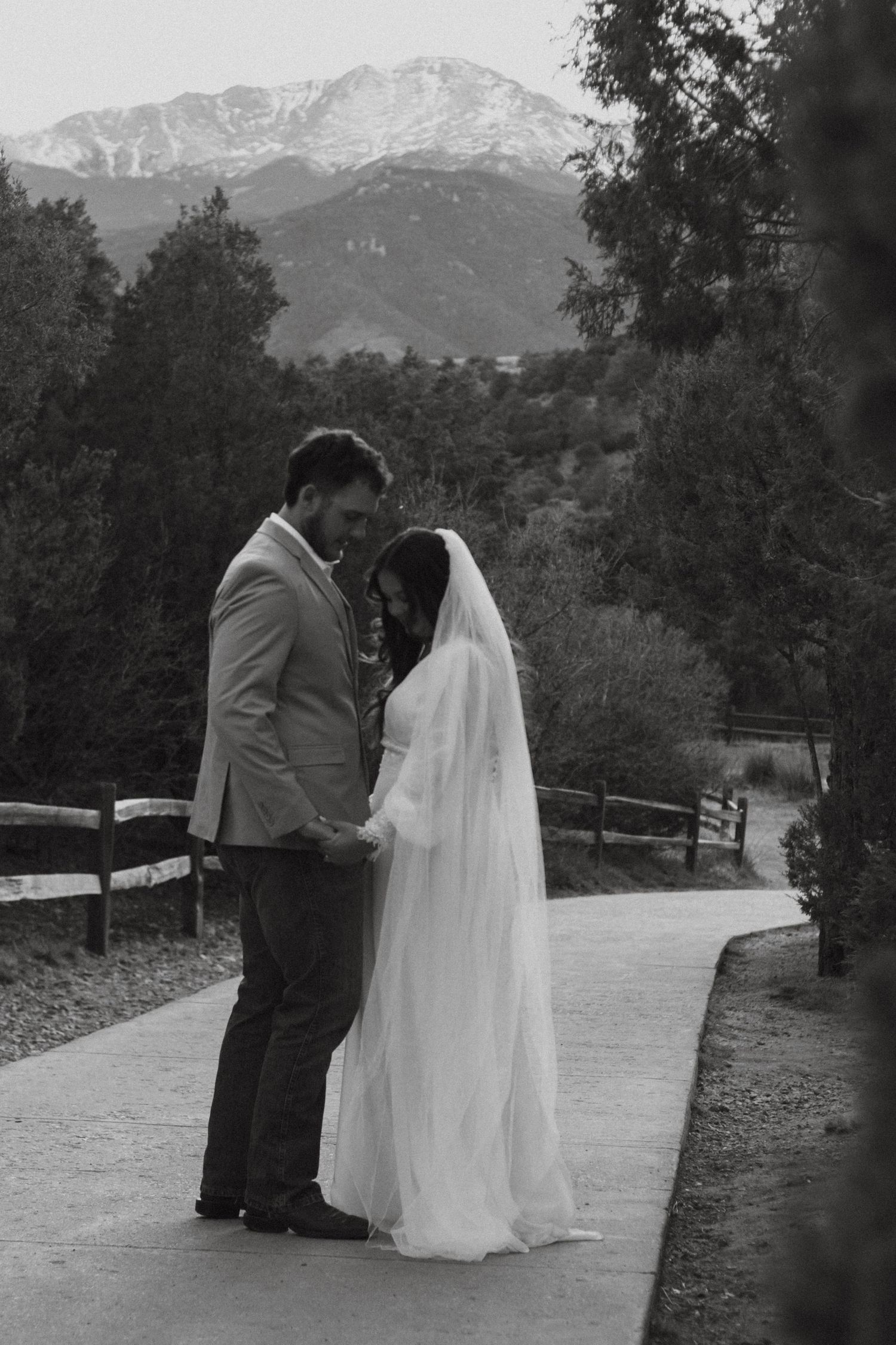 Black and white wedding photo of bride and groom holding hands on a path with mountain backdrop.
