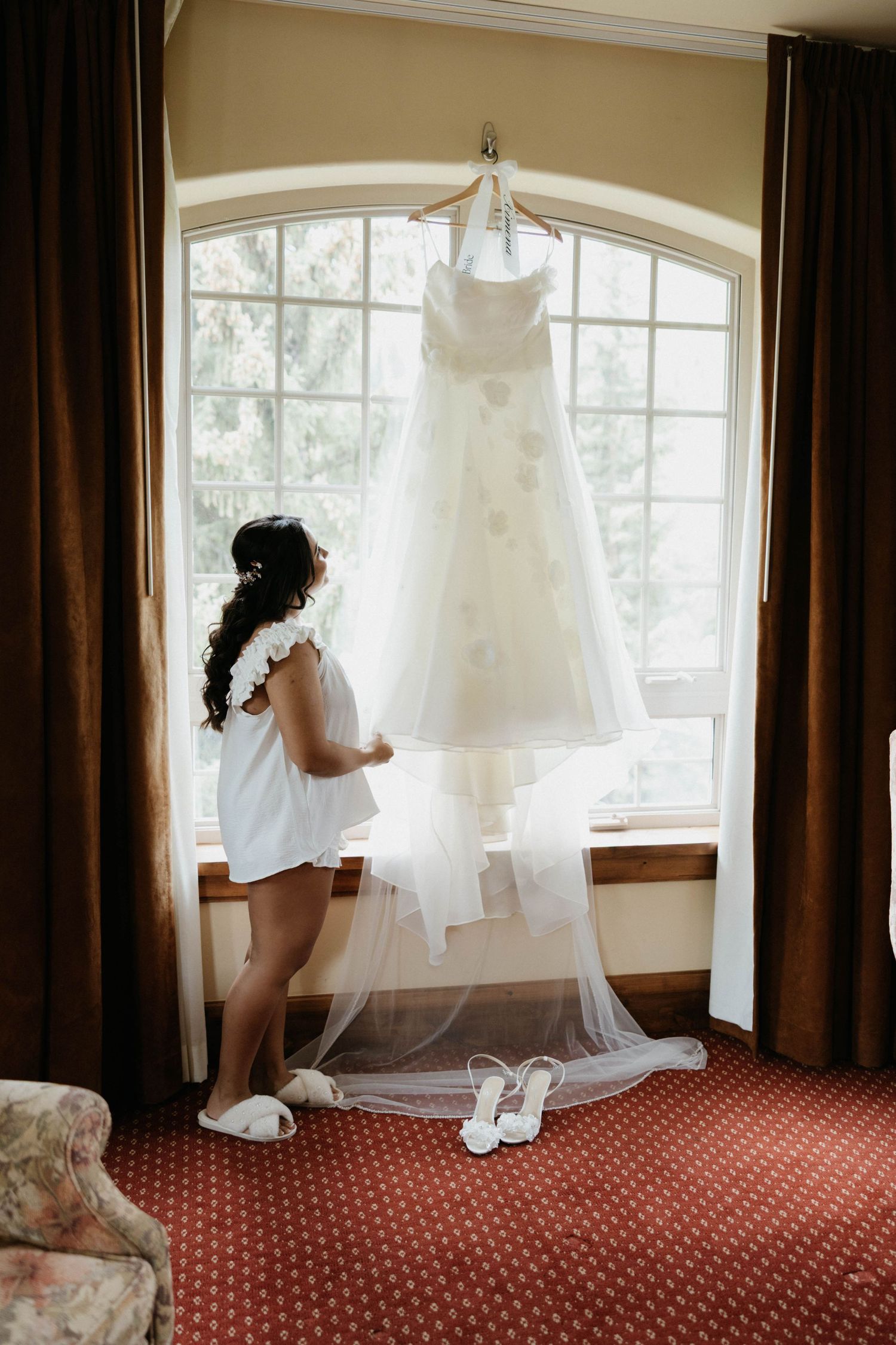 Bride in white robe admiring her wedding gown hanging by a window, with veil and bridal heels displayed below.