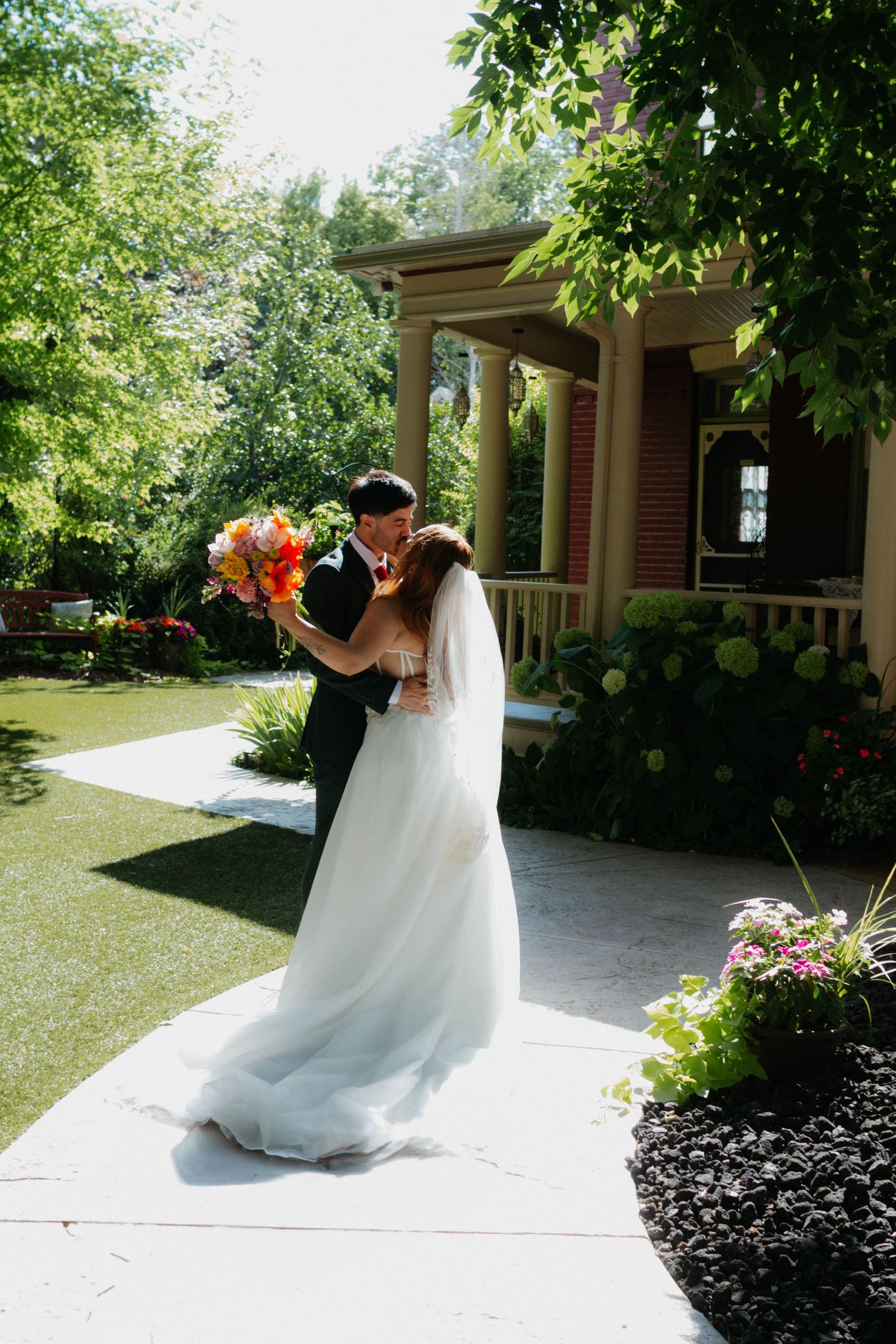 Bride and groom share a romantic kiss on a garden path outside a charming house, with lush greenery and colorful flowers.