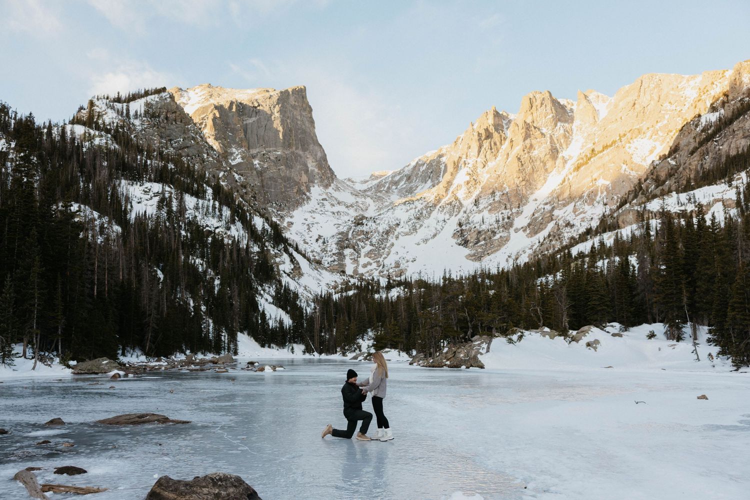 Couple on frozen mountain lake with snow-capped Rocky Mountain peaks and pine trees in winter landscape.