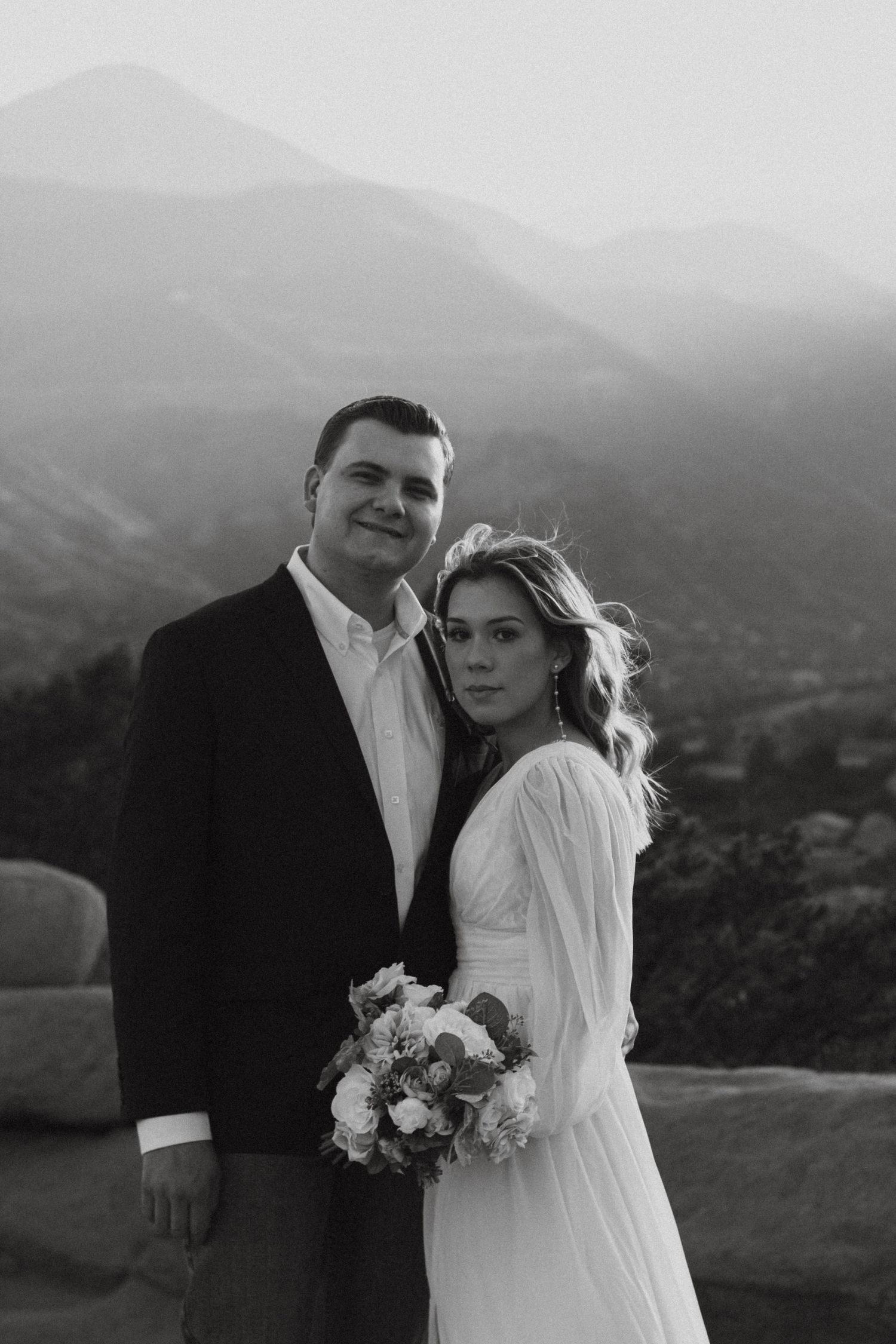 Black and white wedding portrait of bride and groom posing with mountain landscape backdrop.