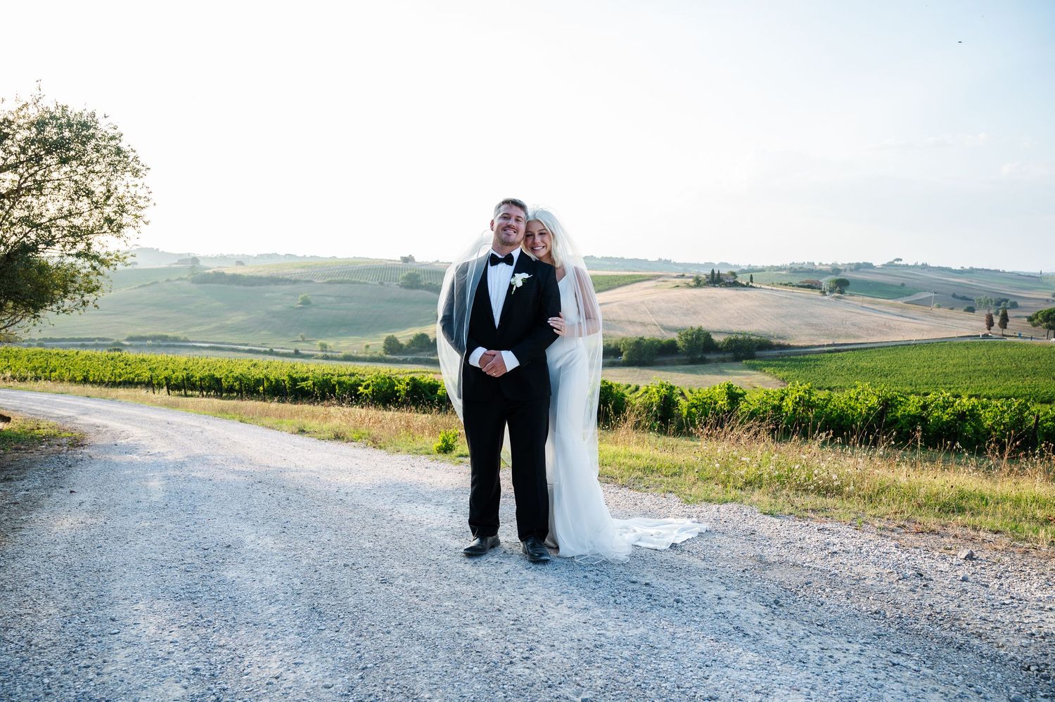 Bride and groom posing on a scenic Tuscany vineyard road at sunset in wedding attire.