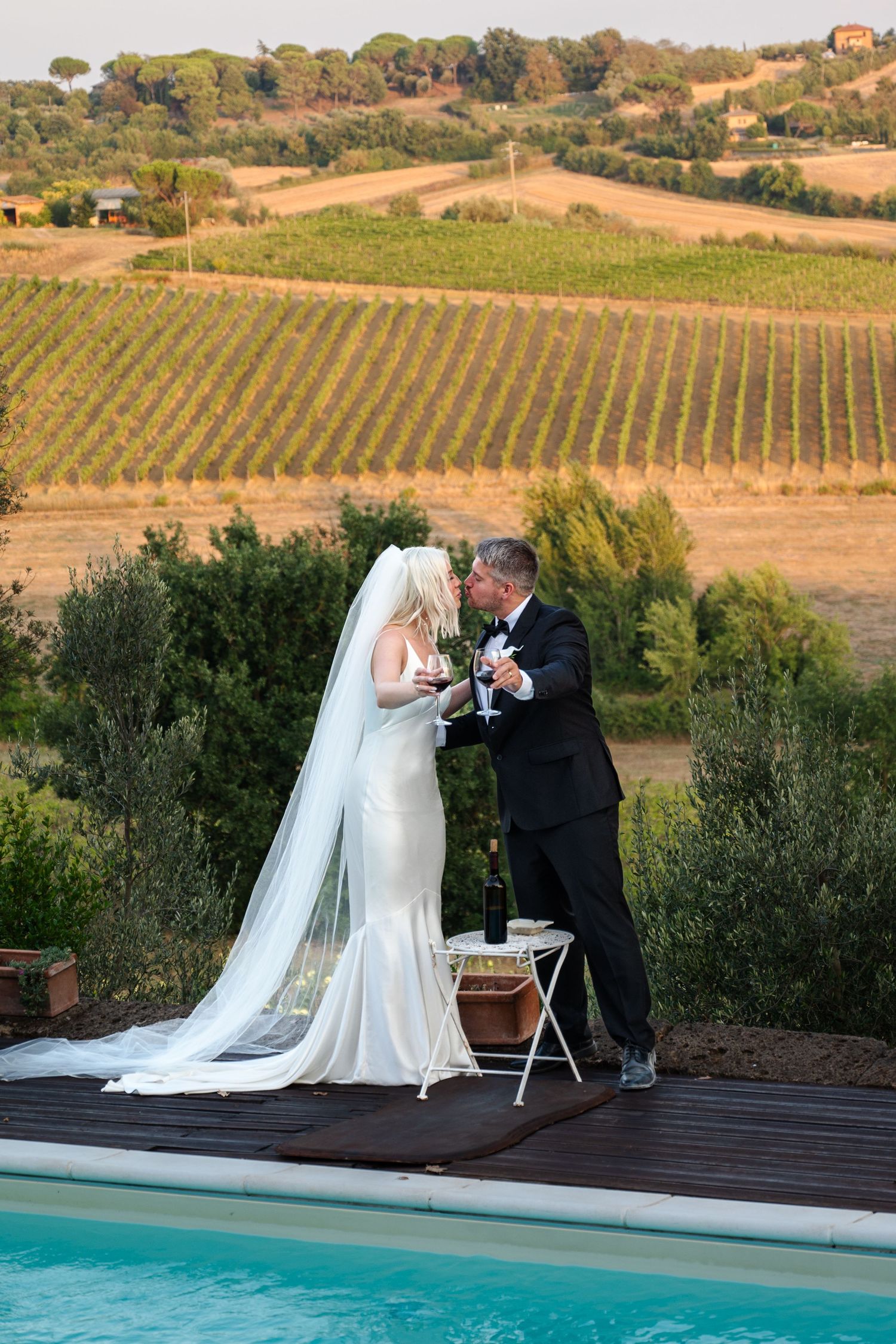 Bride and groom toasting champagne by a pool overlooking stunning Tuscan vineyard at sunset in Tuscany Italy.
