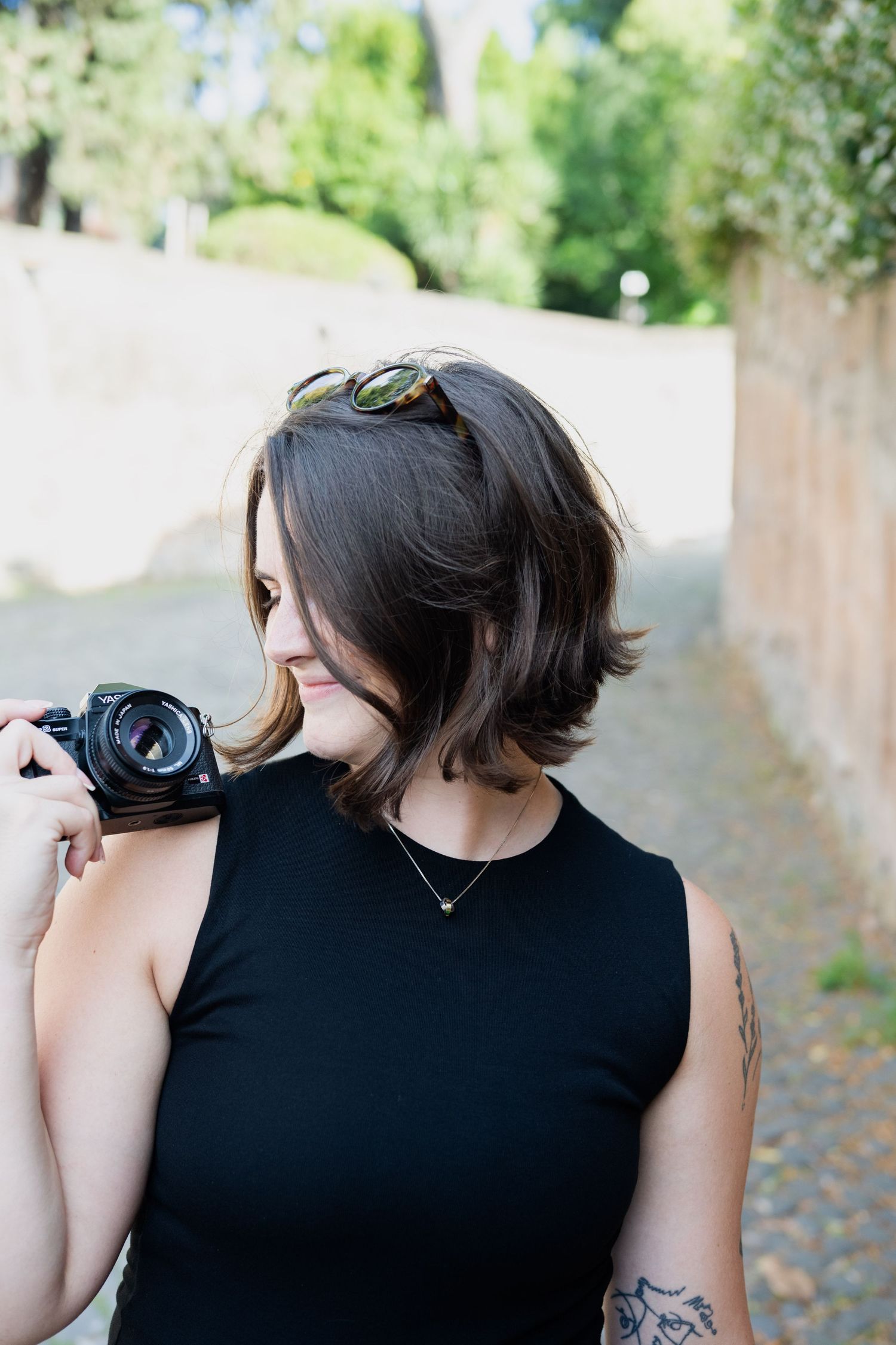 A woman with a short bob hairstyle holds a camera on her shoulder in an outdoor setting.