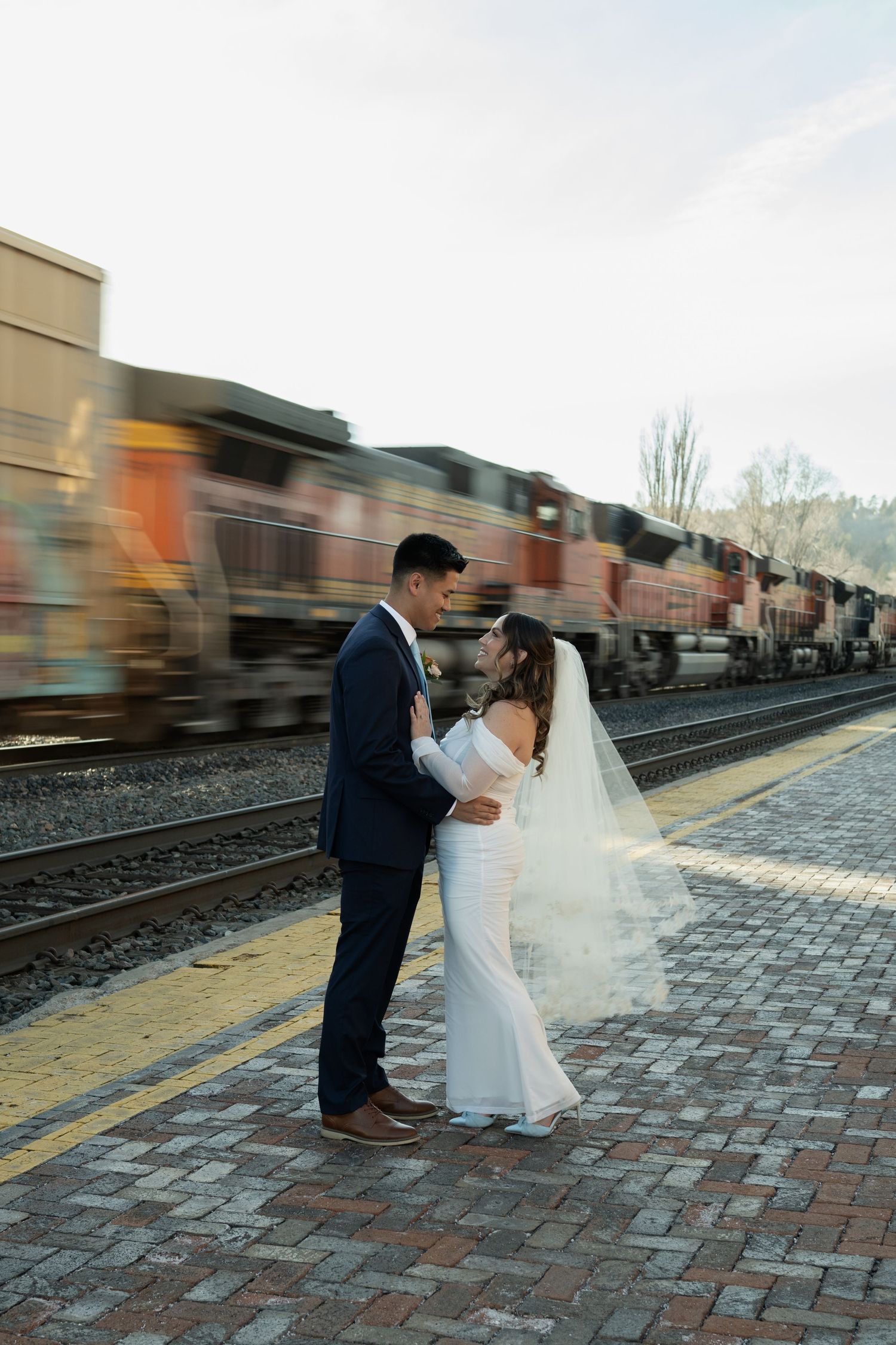 Bride and groom embrace romantically on train platform as BNSF locomotive passes behind them in Flagstaff Arizona.