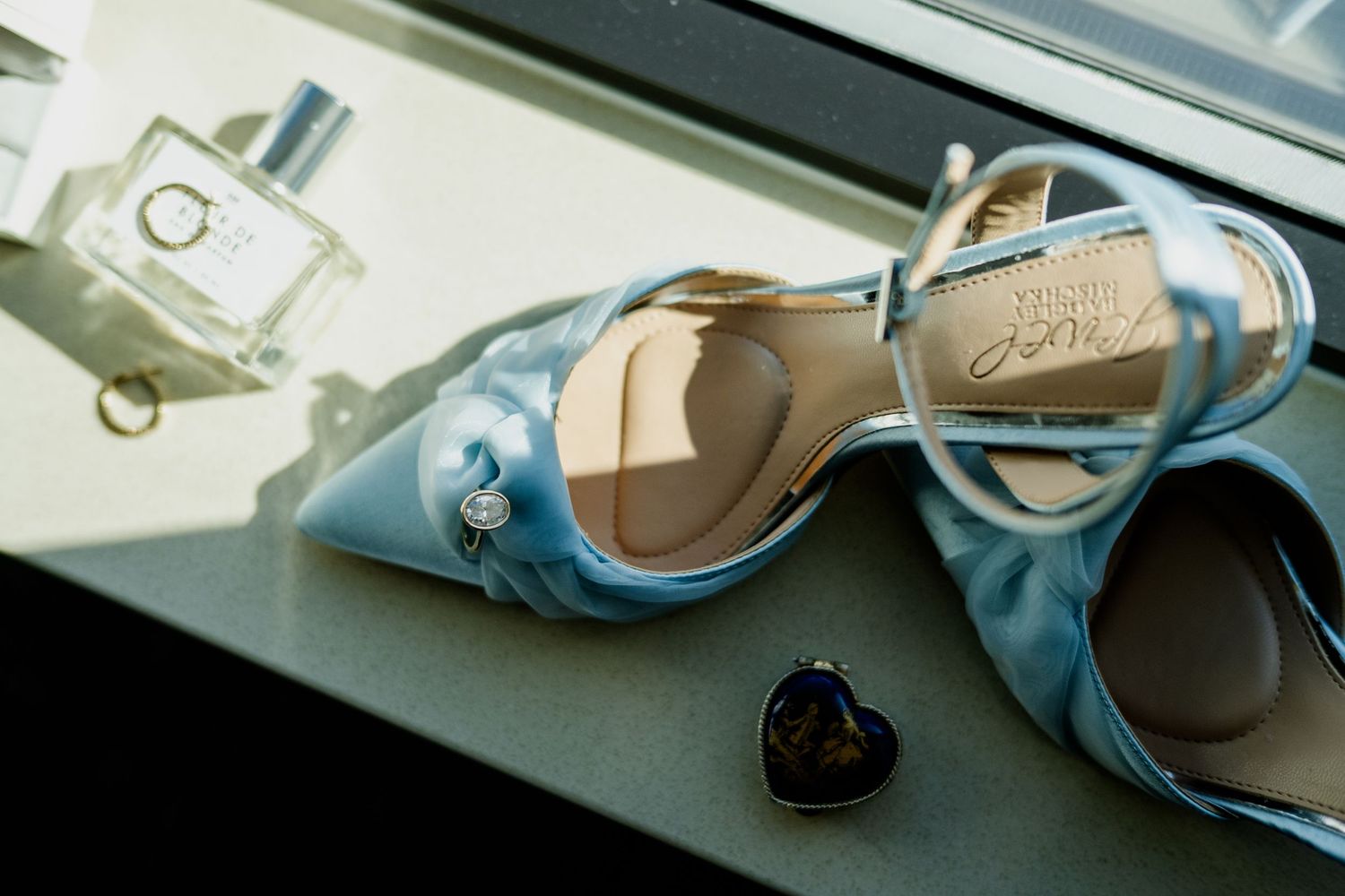 Light blue satin wedding heels with crystal detail displayed on windowsill with perfume bottle and heart jewelry.