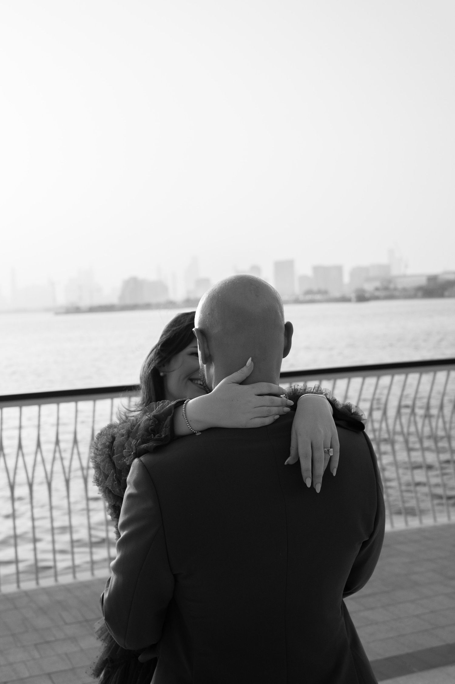 A couple embraces near a waterfront railing with a city skyline in the background in this black and white photo.