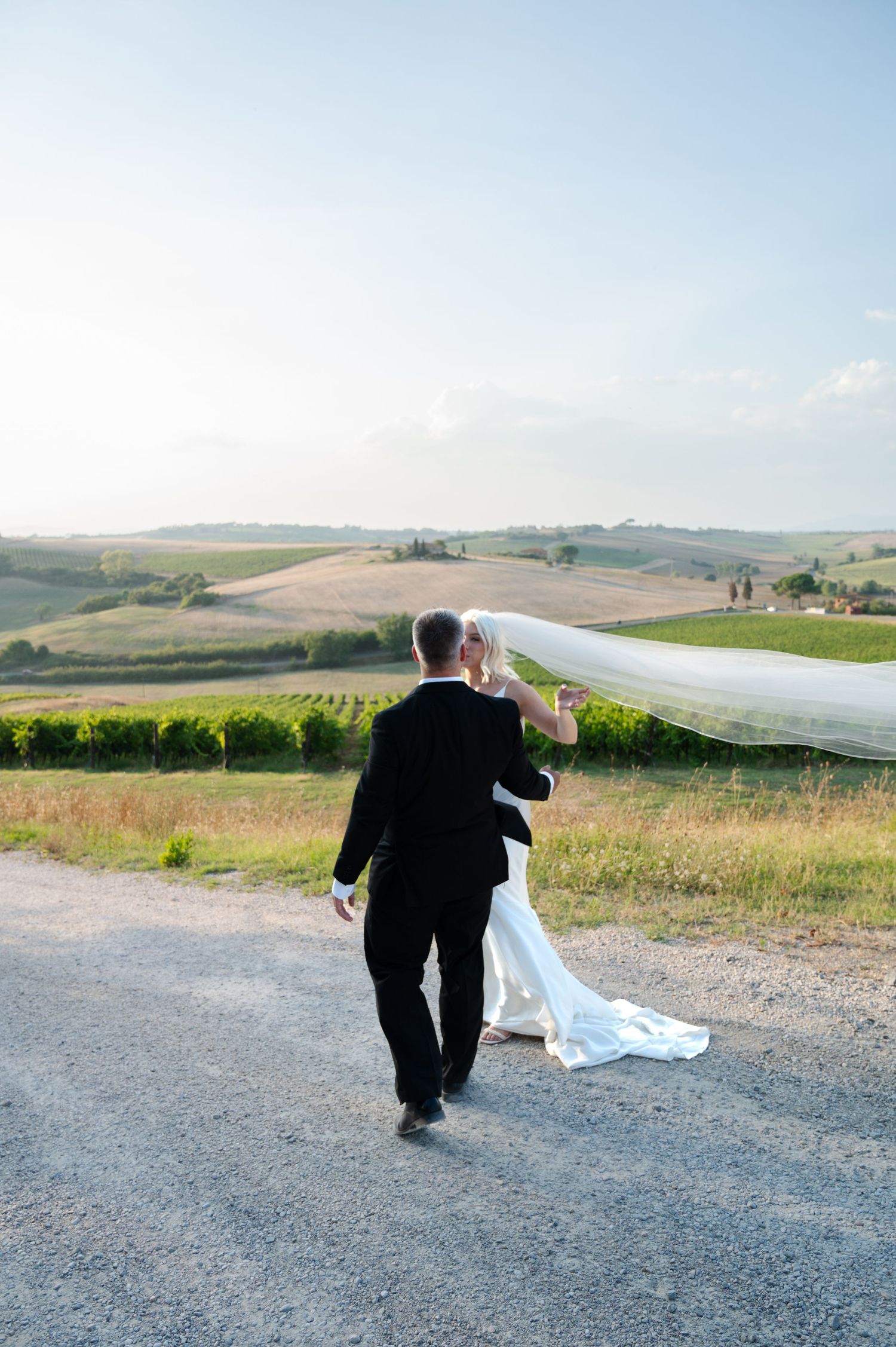 Bride and groom walking along a scenic countryside path, veil flowing in the breeze in Tuscany Italy.