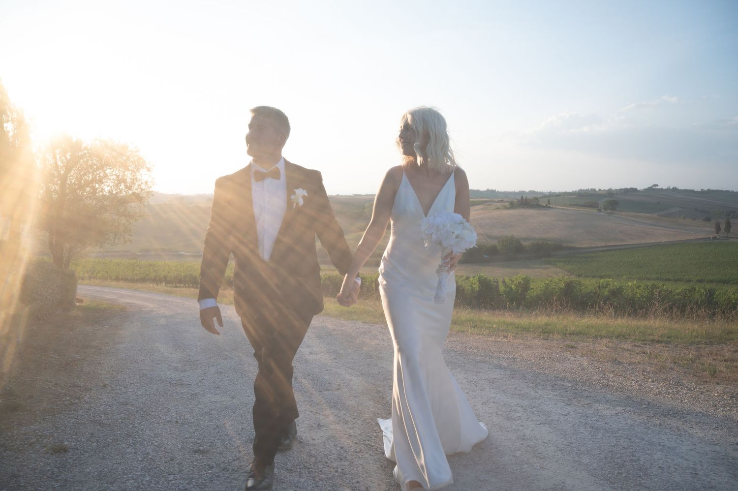 Bride and groom walk hand-in-hand into a golden sunset overlooking sweeping Tuscan countryside vineyards in Tuscany Italy.