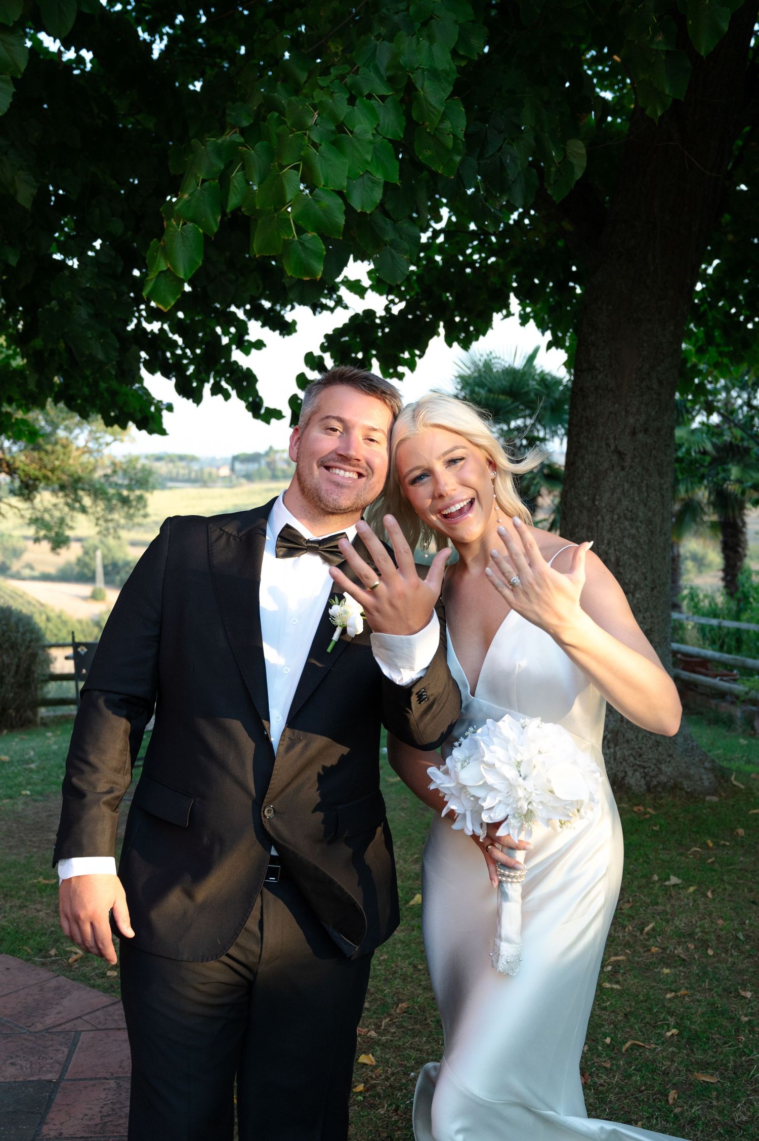 Joyful newlyweds showing off wedding rings while smiling under a tree in the Tuscan countryside in Tuscany Italy.