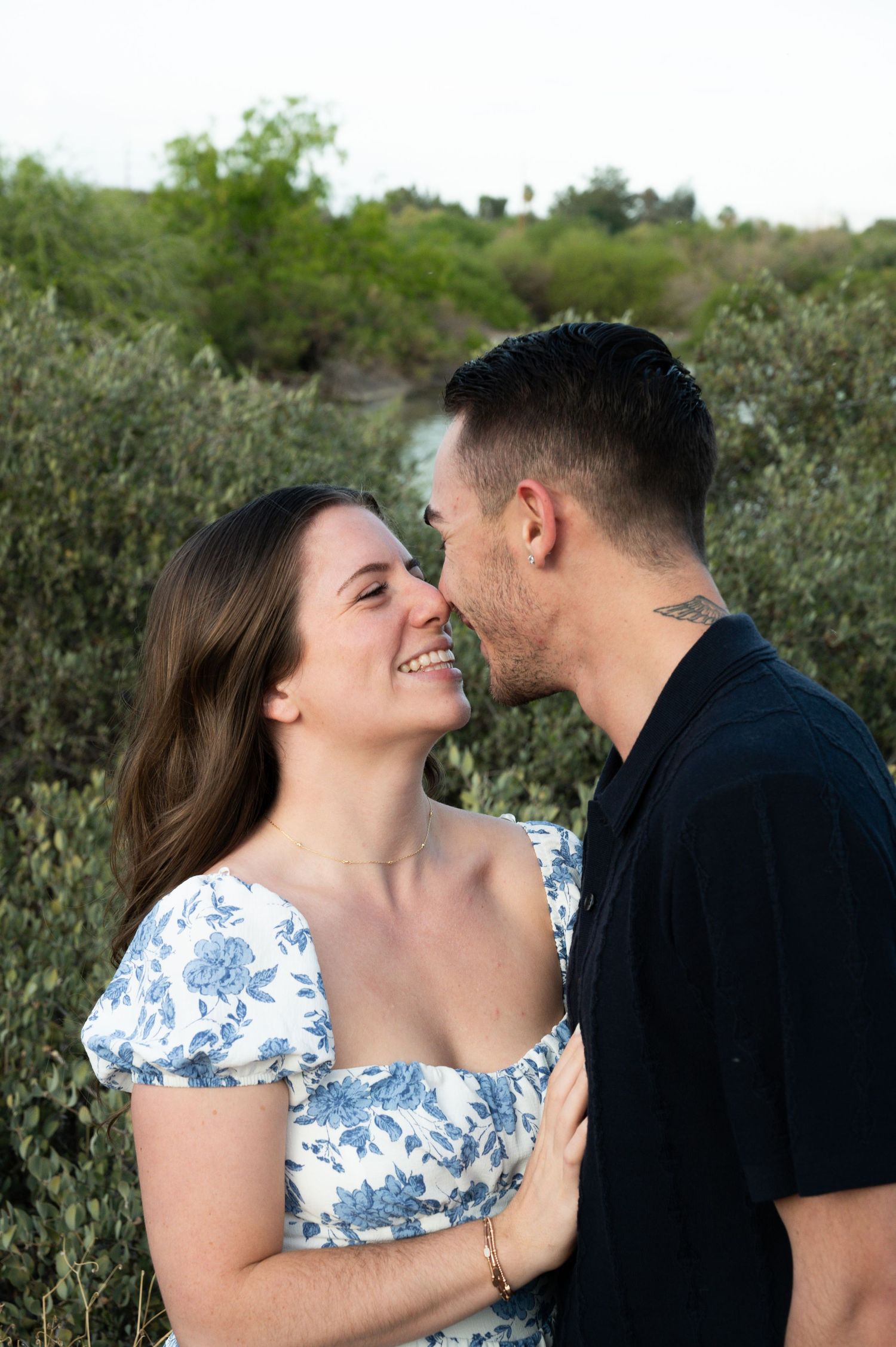 Laughing couple nearly kissing outdoors surrounded by lush green shrubs during their engagement session in gilbert arizona.