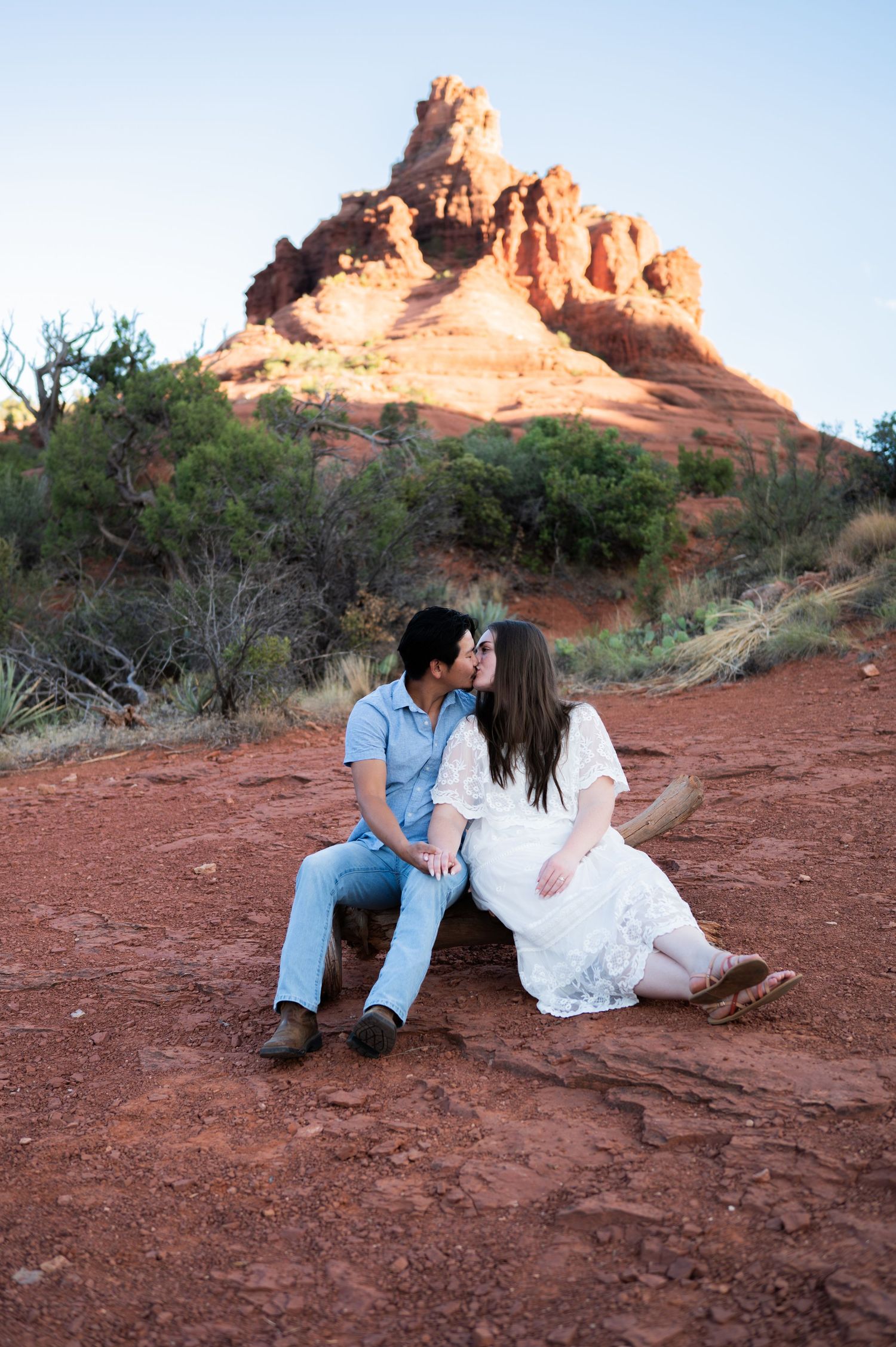 Couple shares a kiss while sitting on red rock ground with Bell Rock formation in background in sedona arizona.