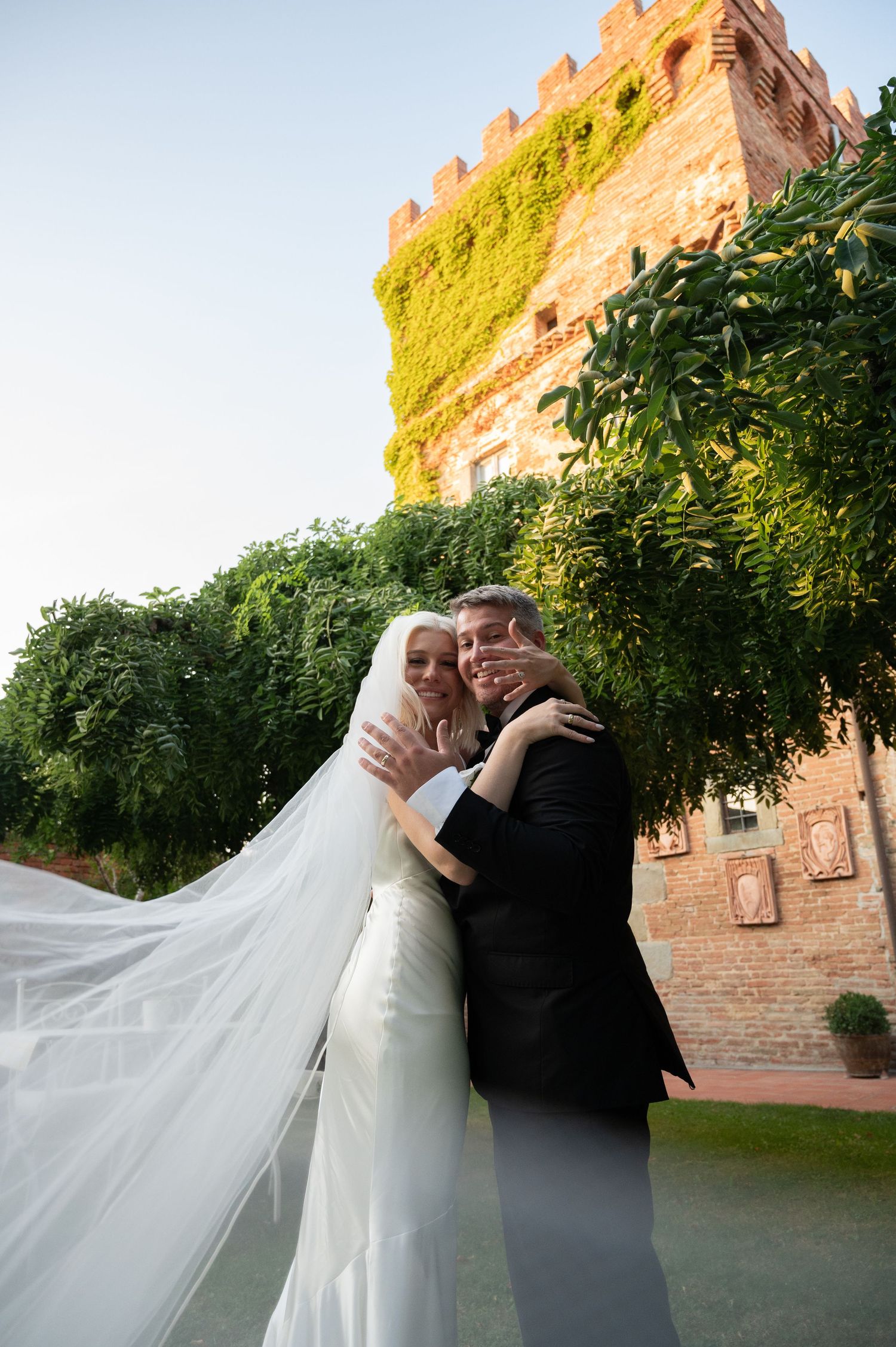 Bride and groom embrace near ivy-covered castle tower on their wedding day, veil flowing in the breeze.