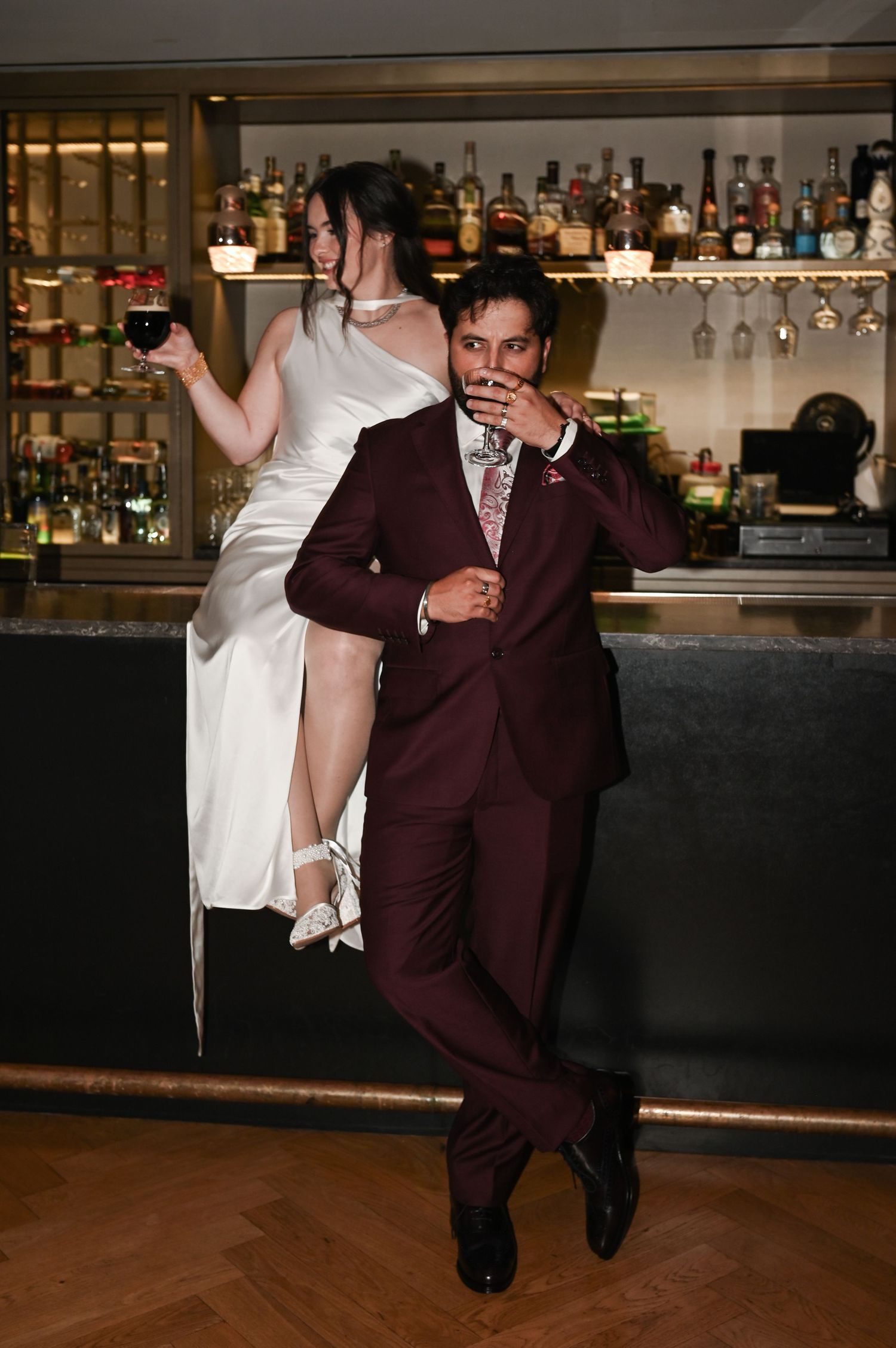 Bride in white gown sits on bar while groom in burgundy suit sips a cocktail at a wedding reception at The Pendry Hotel in Chicago Illinois.