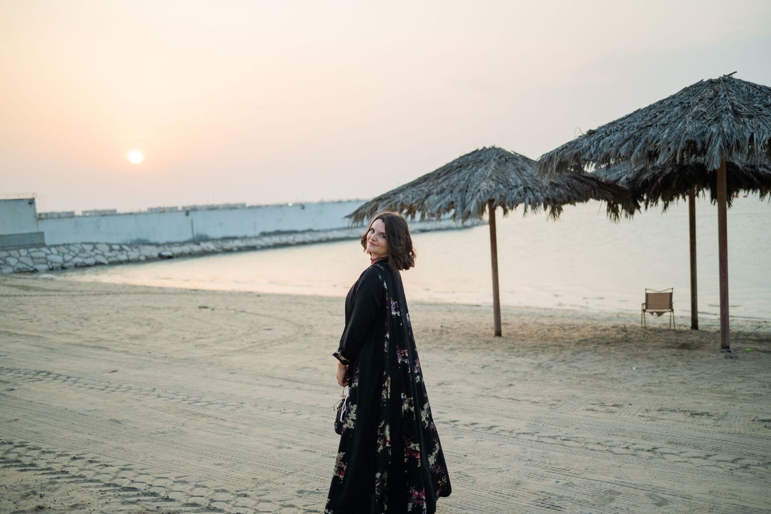 American woman in a black floral Abaya walks on a sandy beach at sunset near straw umbrellas in Jubail Saudi Arabia.