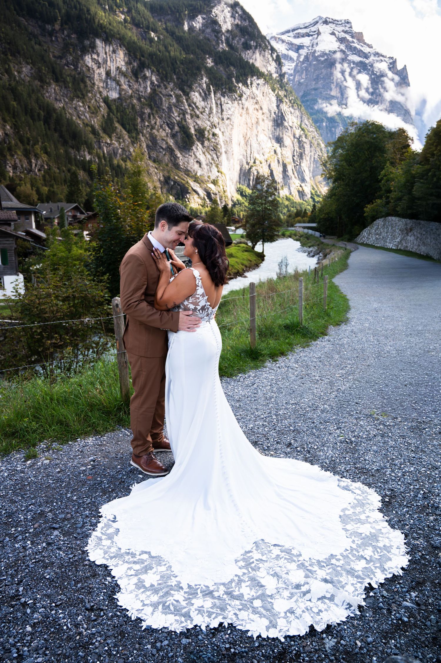 Wedding couple embraces on mountain road with stunning Swiss Alps river valley backdrop at dusk.