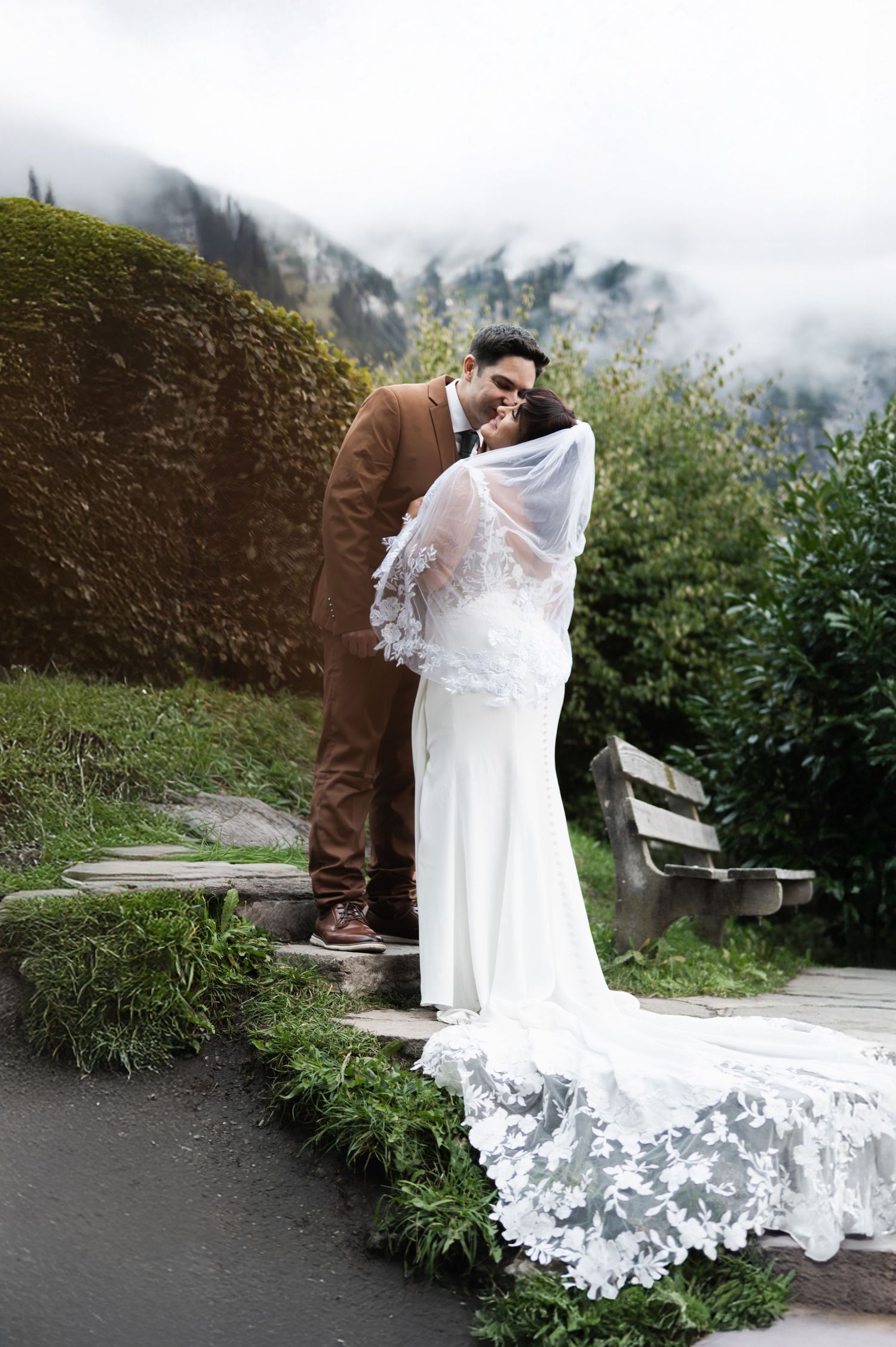 Bride in lace veil and groom in brown suit embracing on stone steps with misty mountains behind in Switzerland.