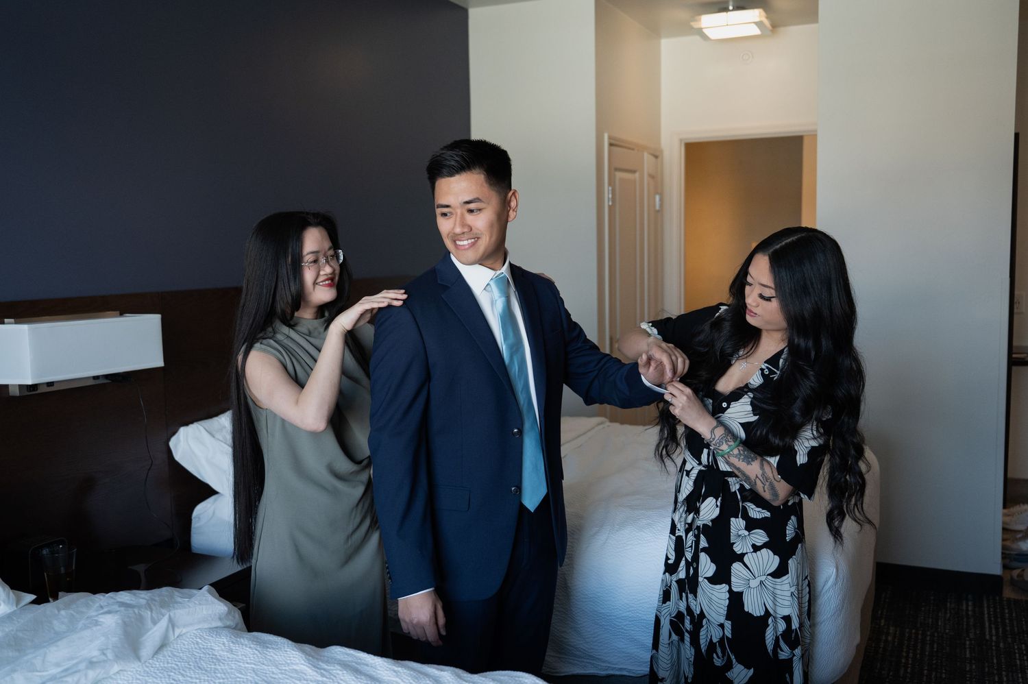 Groom in navy suit being helped into jacket by two women in hotel room before wedding in Flagstaff Arizona.