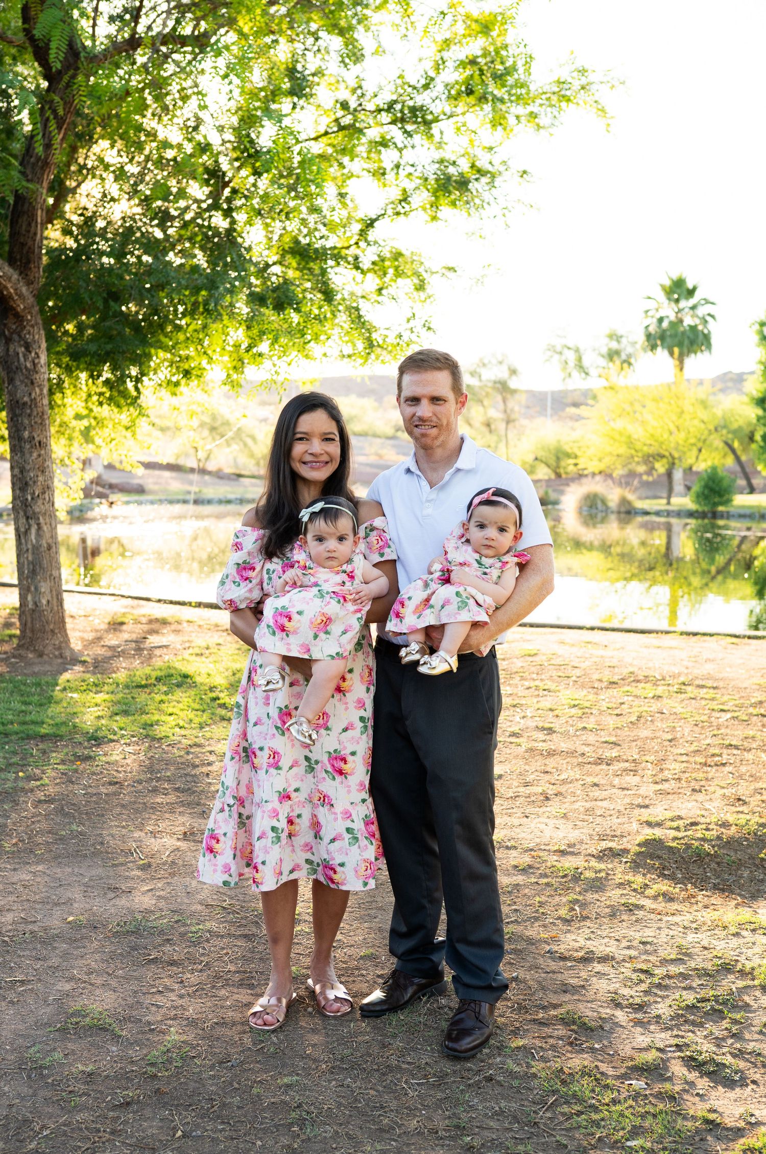 Family of four posing outdoors near a pond, parents holding two baby girls in matching floral dresses on a sunny day.