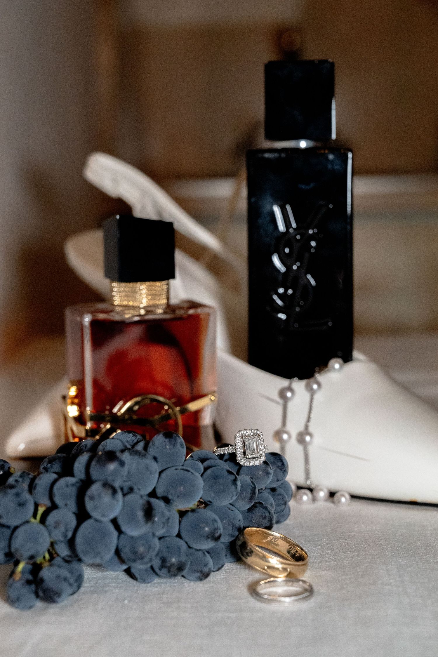 Wedding flat lay with diamond rings, gold band, grapes, YSL perfume bottles, pearl necklace, and white heels in Tuscany Italy.