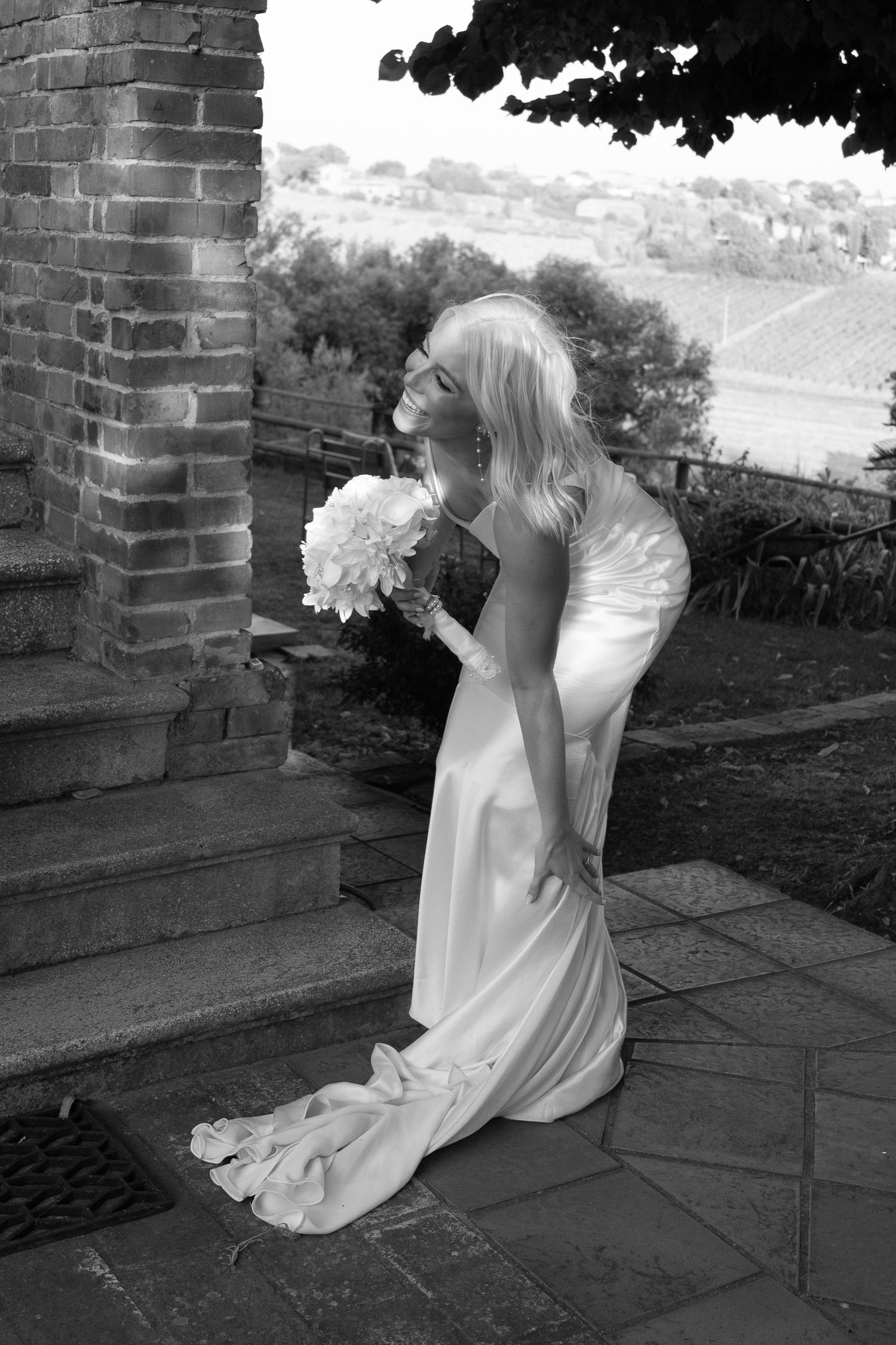 Elegant bride poses on stone steps in a sleek white gown holding a white bouquet in black and white photo in Tuscany Italy.